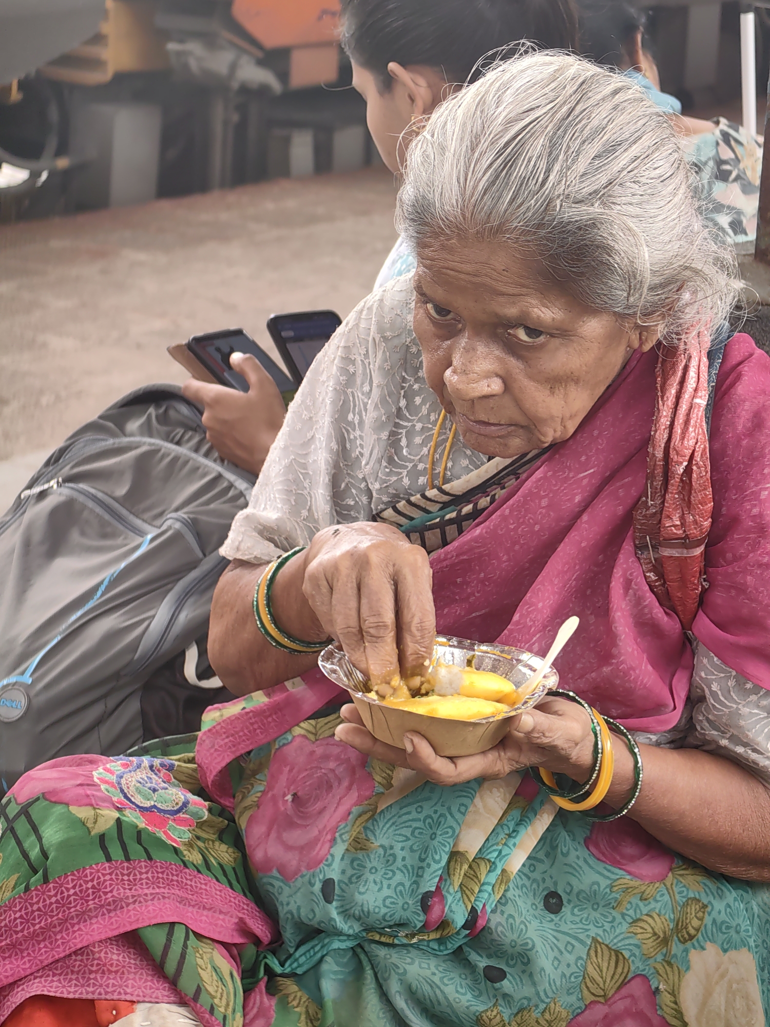 An elderly woman with graying hair, wearing a pink and green floral sari, sits outdoors and eats from a small disposable bowl. Her right hand is in the bowl, picking up food. She is wearing several bangles on her left wrist, including yellow and green ones. A pink scarf is draped around her shoulders, and a grey backpack rests beside her. In the background, another person is partially visible, holding two cell phones. The setting appears to be a public outdoor area, possibly a waiting area or a street scene, given the presence of a backpack and the casual attire of the individuals. The overall scene suggests a moment of everyday life in Itarsi, India.