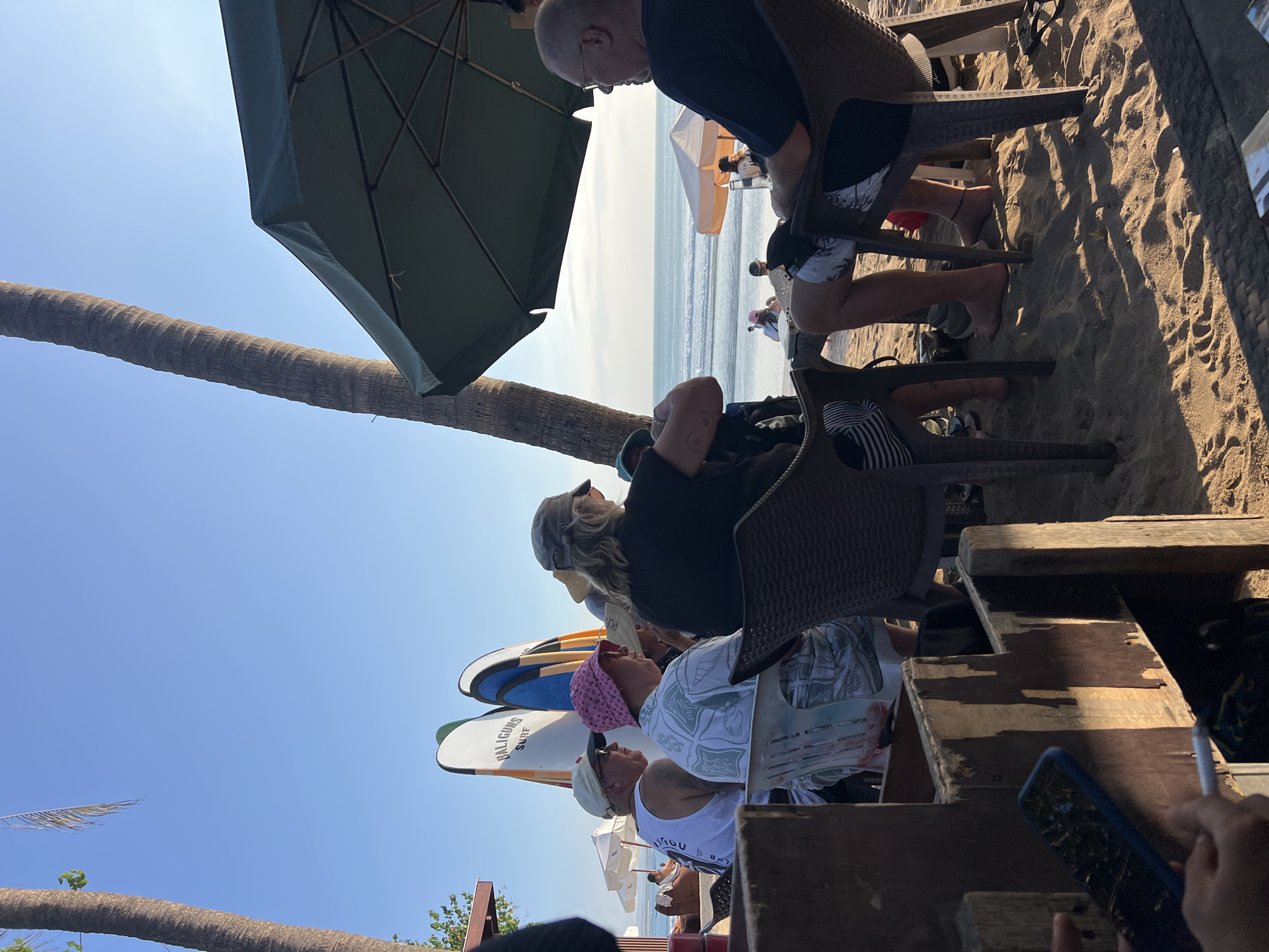 The image depicts a beachside scene in Kuta, Indonesia, likely at a cafe or restaurant. In the foreground, a person's hands are visible holding a mobile phone and a cigarette. Several people are seated at tables on the sandy beach, some under a large green umbrella. To the left, surfboards are propped up against a palm tree. The ocean is visible in the background with gentle waves and a clear blue sky. The overall atmosphere suggests a relaxed day at the beach.
