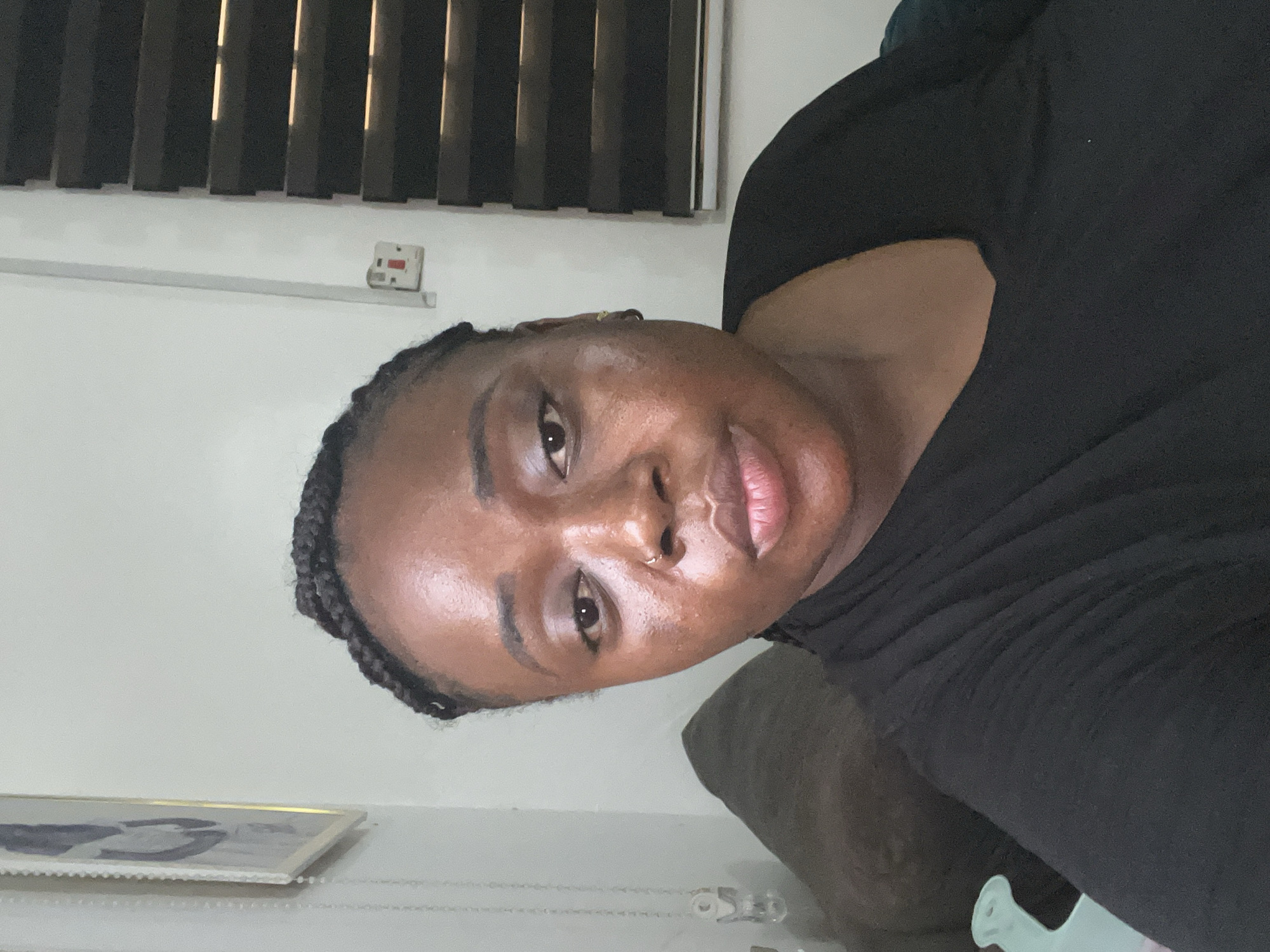 The image is a close-up, vertical selfie of a person indoors, rotated 90 degrees counter-clockwise. The subject is an individual with dark skin and braided hair, wearing a black top. They have a small gold hoop piercing on their right nostril and look directly at the camera with a neutral expression.
The background features dark vertical window blinds at the top, with light filtering through, indicating daytime. Below the blinds, a white wall displays a white electrical outlet or switch with red markings, and a white horizontal rail or strip. A partially visible framed picture, possibly black and white, rests on a surface in the lower left. To the right of the person, a dark piece of furniture, likely a sofa or chair, is visible, along with an unidentifiable light blue object in the bottom right foreground.
The setting is an indoor residential space, situated in Abuja, Nigeria, according to the provided context. No visible text is present in the image.