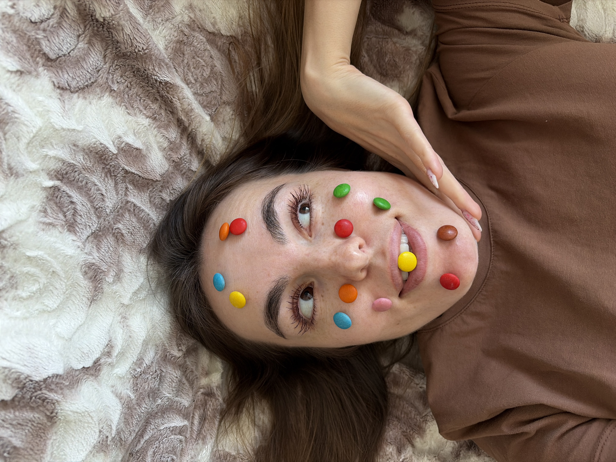 A young woman lies on a beige and brown fluffy blanket wearing a brown top. Various colored candies are stuck to her face, and one yellow candy is between her teeth. Her right hand is near her face, with nails painted with white and pink designs. The location is not evident in the image.