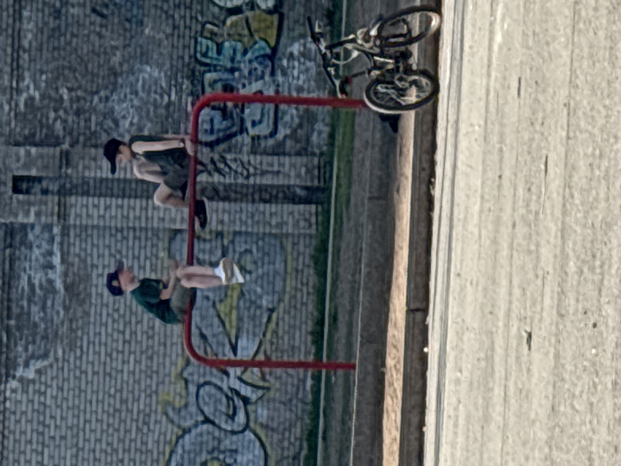 Two people hang on a red horizontal bar on the street in Chaikovsky, Russia. One person wears a dark baseball cap and a dark green T-shirt. The other person wears a dark baseball cap and a sleeveless dark top. A bicycle leans against the red horizontal bar. The background includes a brick wall covered in graffiti and a concrete path.
