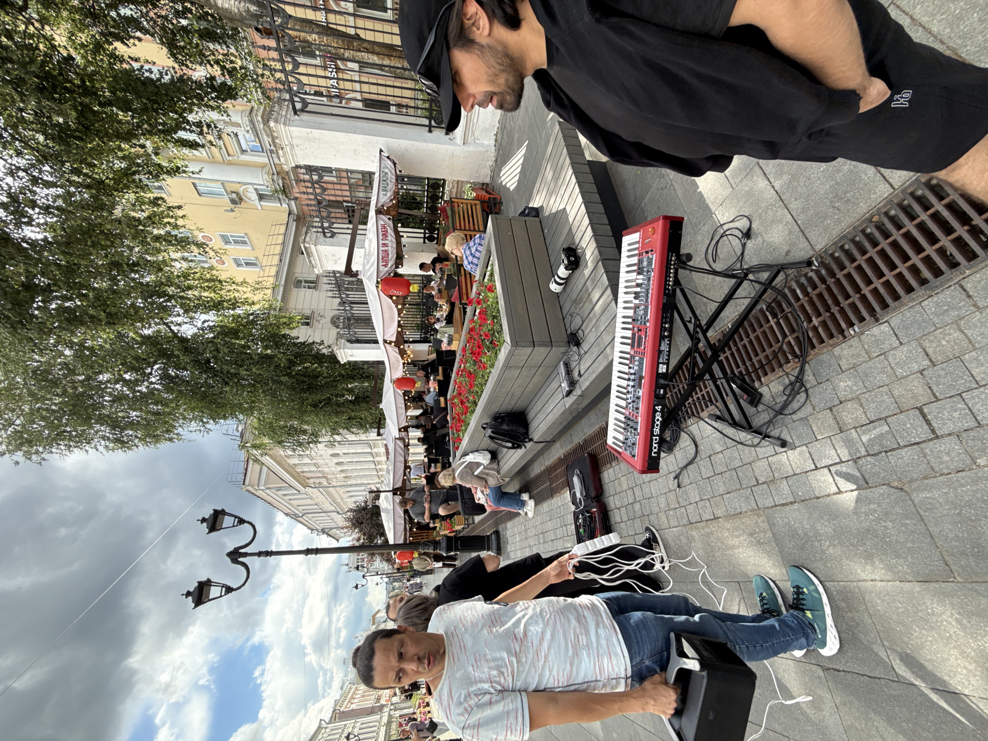 A man in a black cap and black shirt stands in the foreground, looking down. He is holding a black case. Next to him, on the right, is a red keyboard on a stand. Cables are connected to the keyboard. A drain grate is visible on the ground.
In the background, a street scene unfolds with people sitting at tables under umbrellas, seemingly at an outdoor cafe. Red lanterns are hung above the seating area. Buildings with multiple stories line the street. Trees with green foliage are also present. The sky is cloudy.
A woman in a grey sweater and jeans is seated at one of the cafe tables, looking down. Another person in a blue and white checkered shirt is also visible. A camera is placed on a wooden surface near the keyboard. Red flowers are planted in a raised bed.
The scene appears to be taking place during the daytime. The overall atmosphere suggests a casual street performance or a lively outdoor public space. There is visible text on the keyboard which reads "nord stage 4".