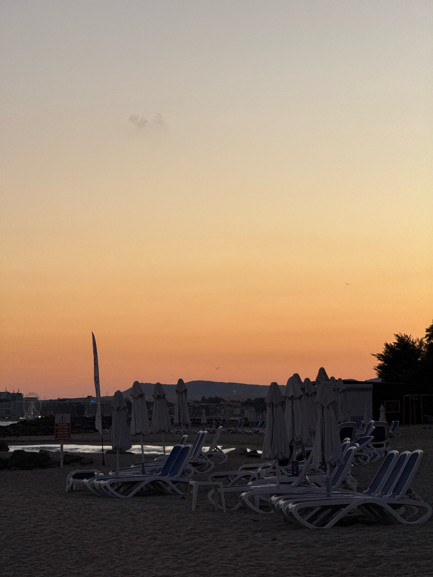 The image depicts a beach scene in Sveti Vlas, Bulgaria, during sunset. The sky is a gradient of soft orange and yellow hues, transitioning to a pale gray at the top. A few wispy clouds are visible in the upper portion of the sky.
In the foreground, rows of empty lounge chairs are arranged on the sandy beach. Most of the chairs are white with blue accents. Several beach umbrellas, closed and covered, are standing upright behind the lounge chairs, indicating the end of the day or preparation for closure.
Further back, the coastline is visible with a body of water, possibly the Black Sea, reflecting the sky's colors. A few dark shapes, possibly rocks or a breakwater, are present near the water's edge. In the distance, a Ferris wheel and buildings suggest a populated area or resort town. Silhouetted trees and hills form the horizon line on the right and left sides of the image.
A tall, thin pole, possibly a flagpole or part of a structure, stands prominently in the middle ground. A red and white sign with text is also visible near the water, but the text is illegible. A few birds are seen in flight against the sky. The overall atmosphere is serene and quiet, typical of a beach at dusk. No people are visible in the frame.