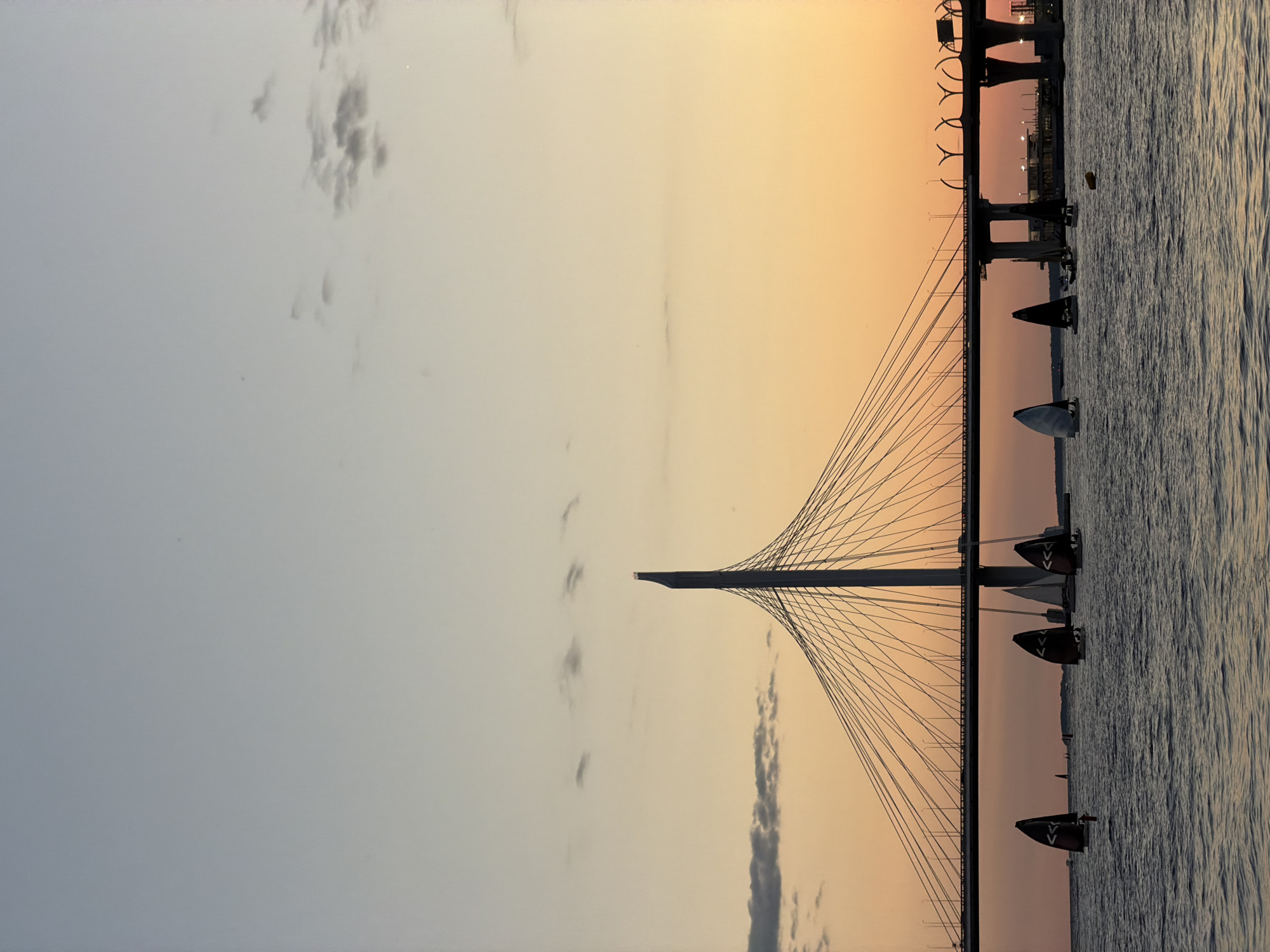 This image captures a sunset over the Gulf of Finland in Saint Petersburg, Russia, with the Western High-Speed Diameter cable-stayed bridge prominently featured. Numerous sailboats with dark sails are visible on the water, appearing to be engaged in a race or leisure sailing. The sky transitions from a pale blue at the top to a warm orange and yellow near the horizon, with a few scattered clouds. The bridge's distinctive pylon and cables are silhouetted against the colorful sky. The water appears calm with gentle ripples. The overall scene evokes a serene and picturesque atmosphere of a coastal city at dusk.
