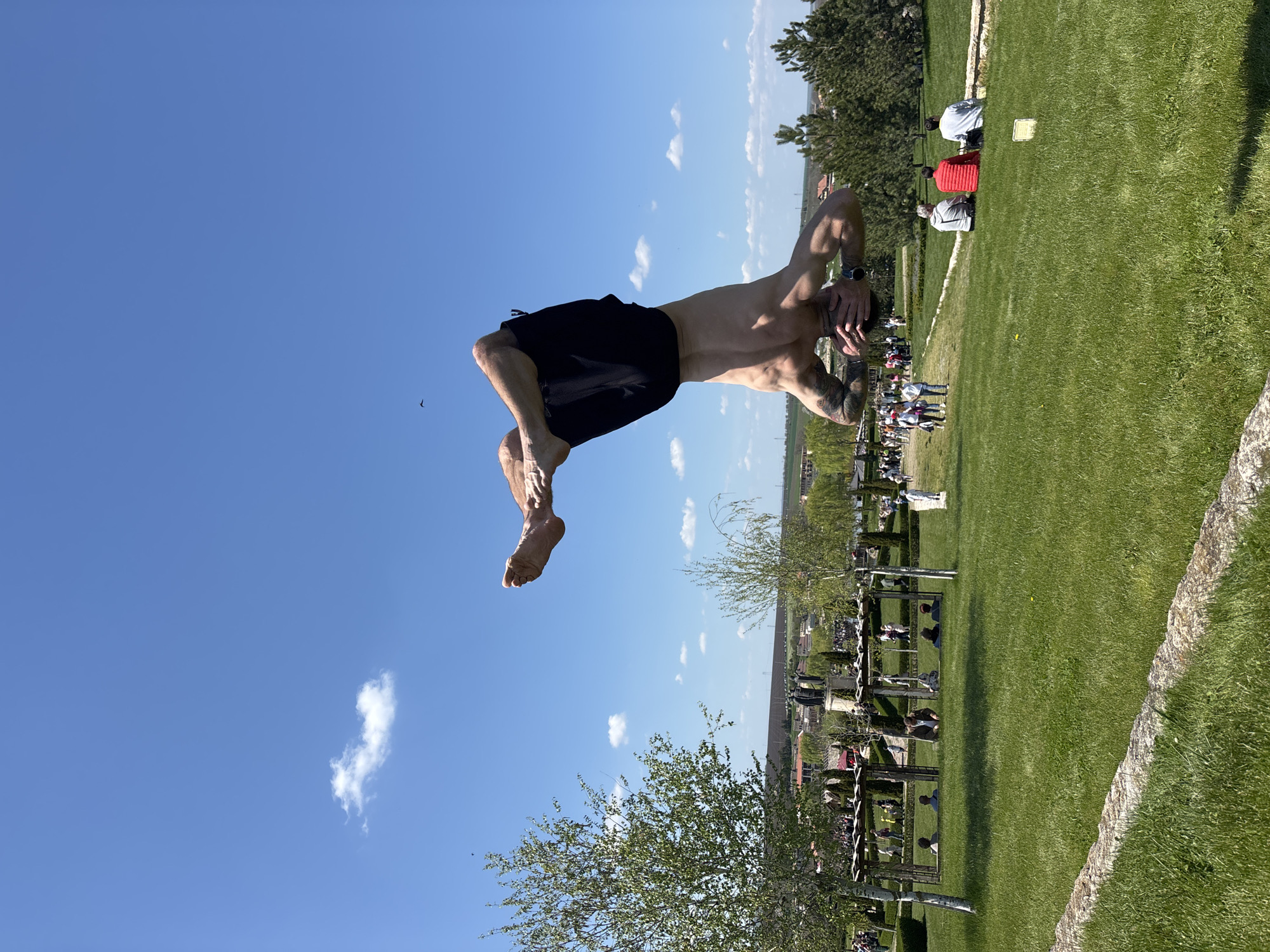 A man is captured mid-air while performing a backflip, appearing to be against a clear blue sky with a single cloud and a bird. He is wearing dark shorts, has a watch on his left wrist, and tattoos on his left arm. He is oriented horizontally in the frame. Below him is a verdant grassy area where several individuals are seated under structures, possibly arbors. In the background, there appears to be a town or city, suggesting the event is taking place in a park or outdoor gathering space in an urban area. The location is Unknown, Unknown.