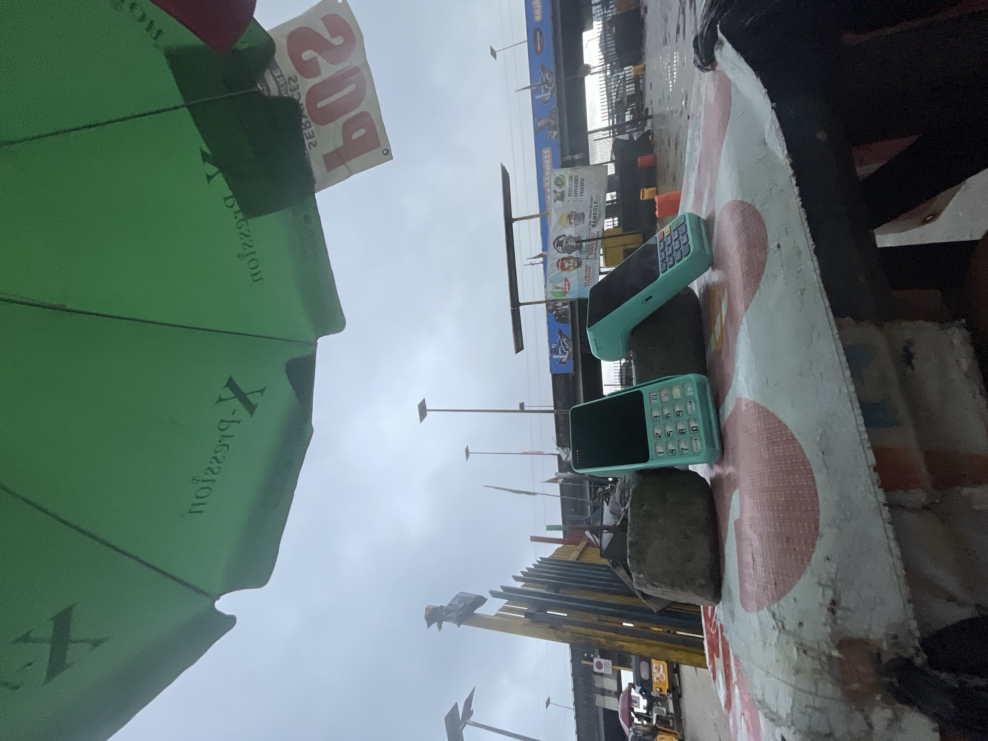 In Ajah, Nigeria, an outdoor scene unfolds under an overcast sky, likely during a period of rain, given the wet ground. In the foreground, two turquoise-colored mobile payment terminals (POS machines) are placed on a weathered, patterned surface that appears to be the top of a makeshift table or cart. One POS machine is upright, while the other is tilted. These are stabilized by two large, irregular gray stones.
Above the foreground objects, a large green umbrella shields the view, its underside displaying text, partially obscured. A white sign with red lettering, reading "609" in reverse, is attached to the umbrella or a nearby structure.
In the mid-ground, a yellow and black striped fence-like structure stands tall, with a black plastic bag tied to its top. Beyond this, a bridge or overpass dominates the background, with various banners and signage displayed on it. These banners feature images of people, political party logos (including APC), and text such as "DEDICATION EXPERIENCE PROGRESS" and "TINU U BANKOLE." Several flags are also visible, along with streetlights.
To the left, a small group of vehicles, including what appears to be a yellow motorized tricycle (Keke Napep), are parked or in motion on a wet street. The overall impression is a busy urban environment, possibly a market or transportation hub, with commercial activities evident.