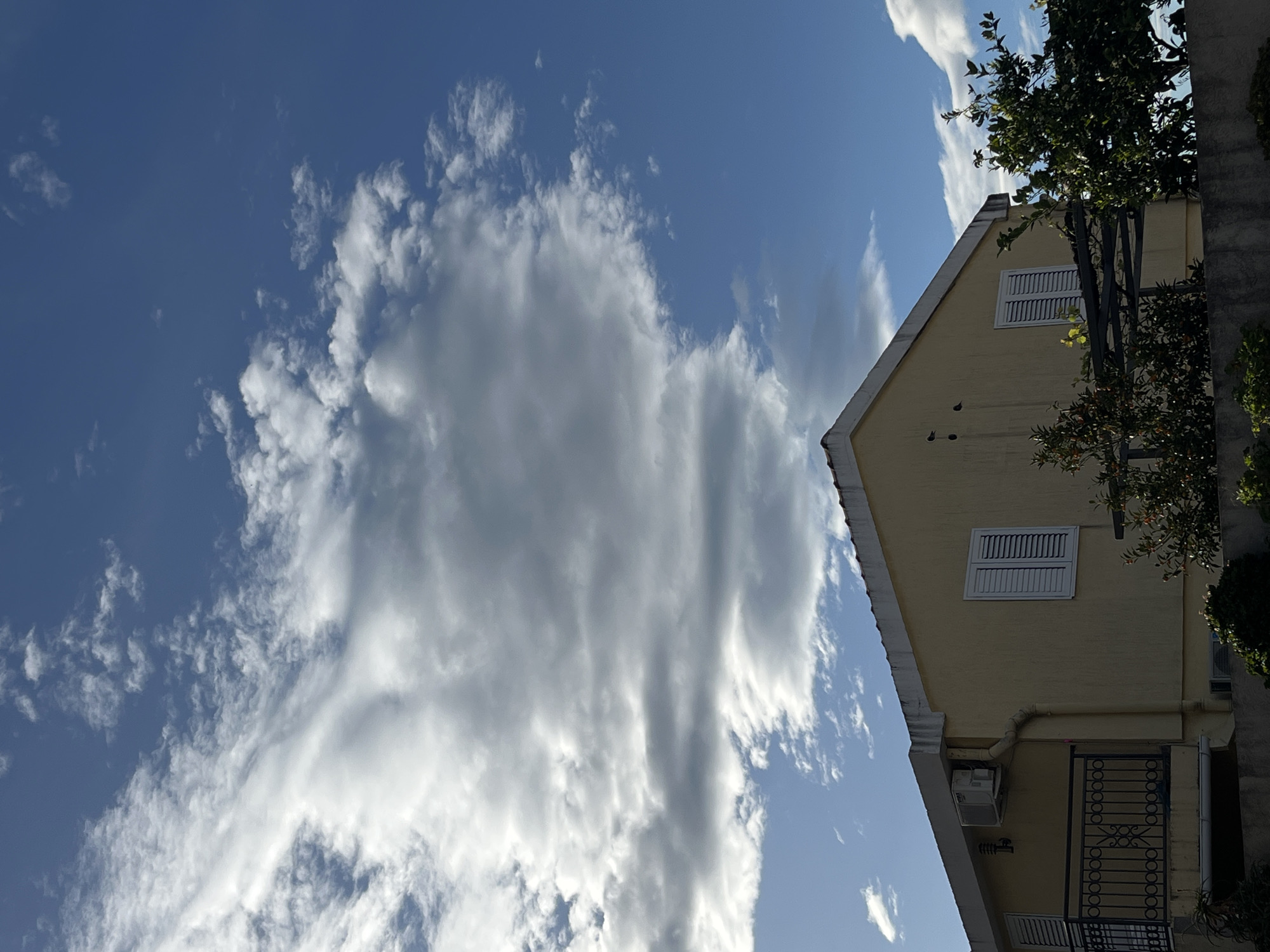 The image shows a light yellow two-story building with white window shutters located in Bečići, Montenegro. The building has a small balcony with a dark metal railing on the first floor. An air conditioning unit is visible on the exterior wall. Part of a tree with green leaves and orange berries is in front of the building. The sky is blue with large, puffy white clouds.