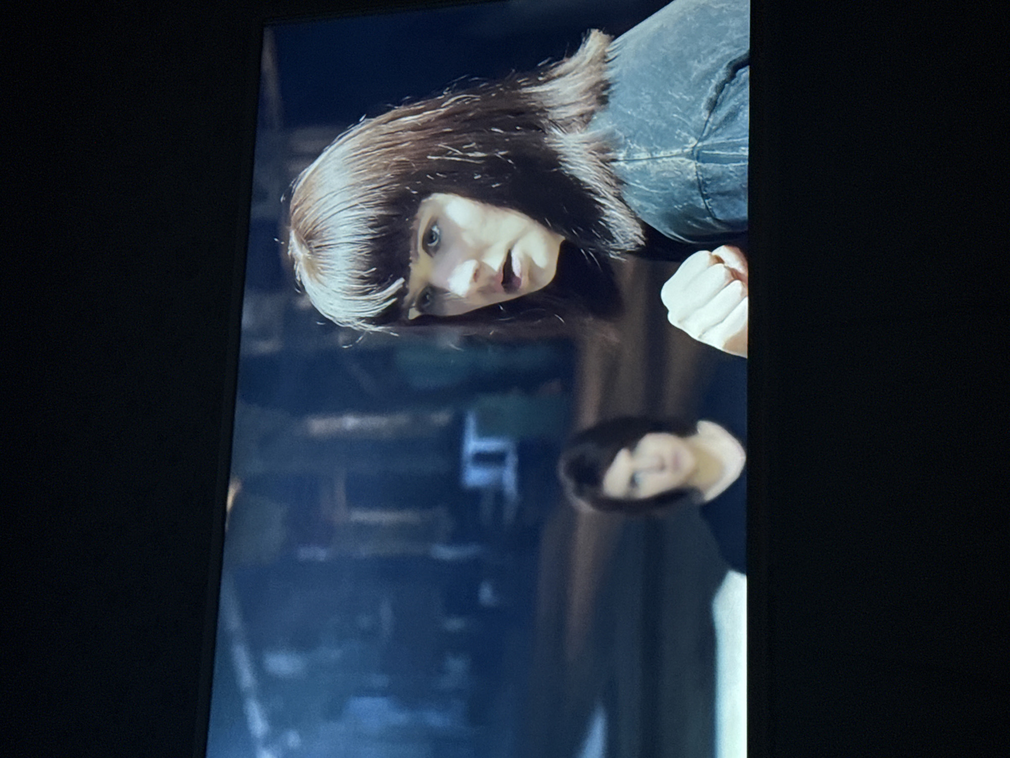 A woman with dark brown hair and bangs is looking intently toward the camera. She has a clenched fist near her face and is wearing a dark jacket. A reflection of another woman with similar dark hair is visible in the background, along with other blurry background elements. The location is unknown, including if it is within the city of Mytishchi, Russia.