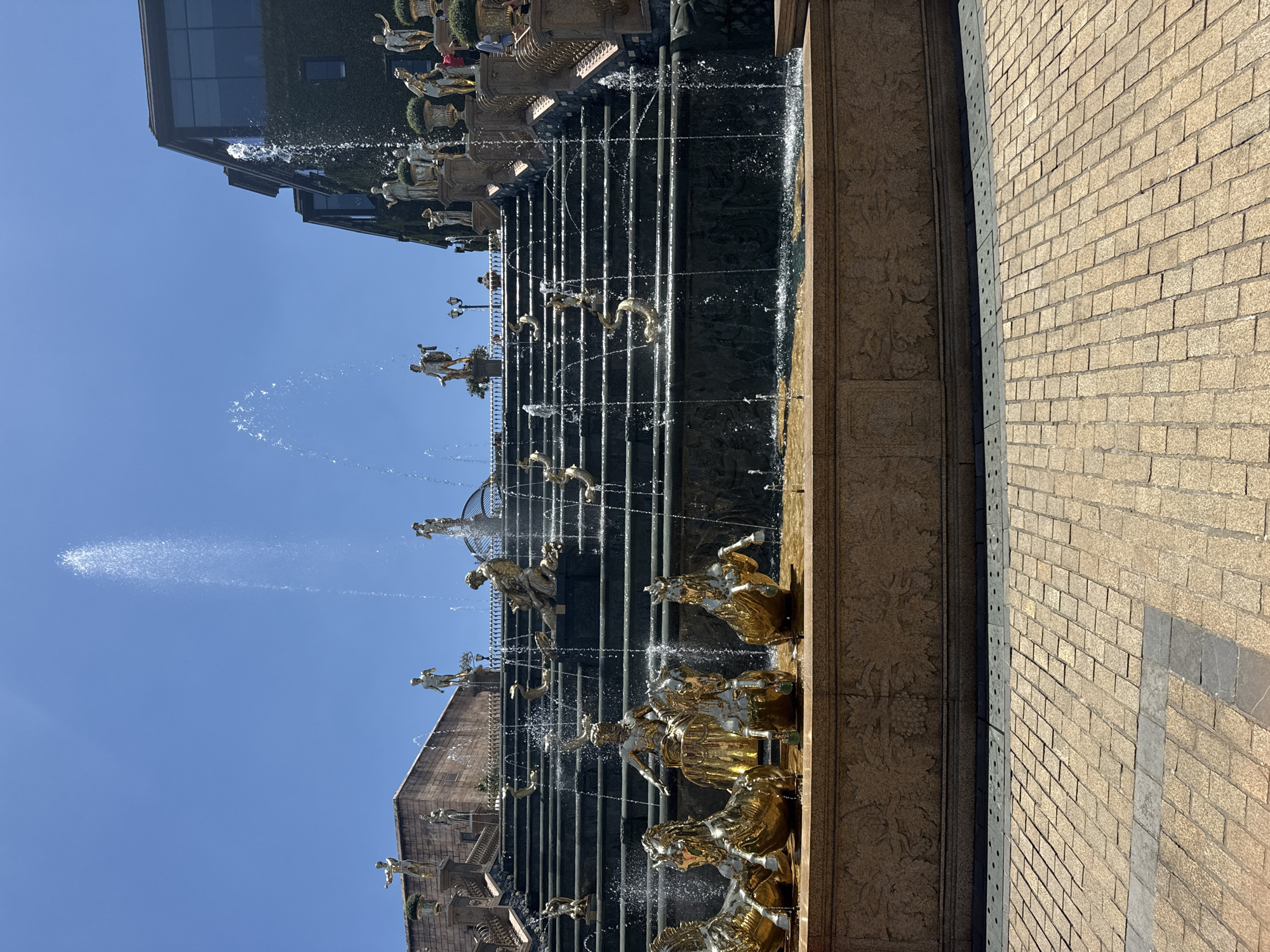 The image shows a grand fountain in Đà Nẵng, Vietnam, likely near the city center. The fountain is multi-tiered with golden statues of figures and animals. Water cascades down multiple levels with visible spray. A building with dark windows and some green foliage is in the background. A brick pathway with a dark stripe is to the right. The sky is clear and blue.