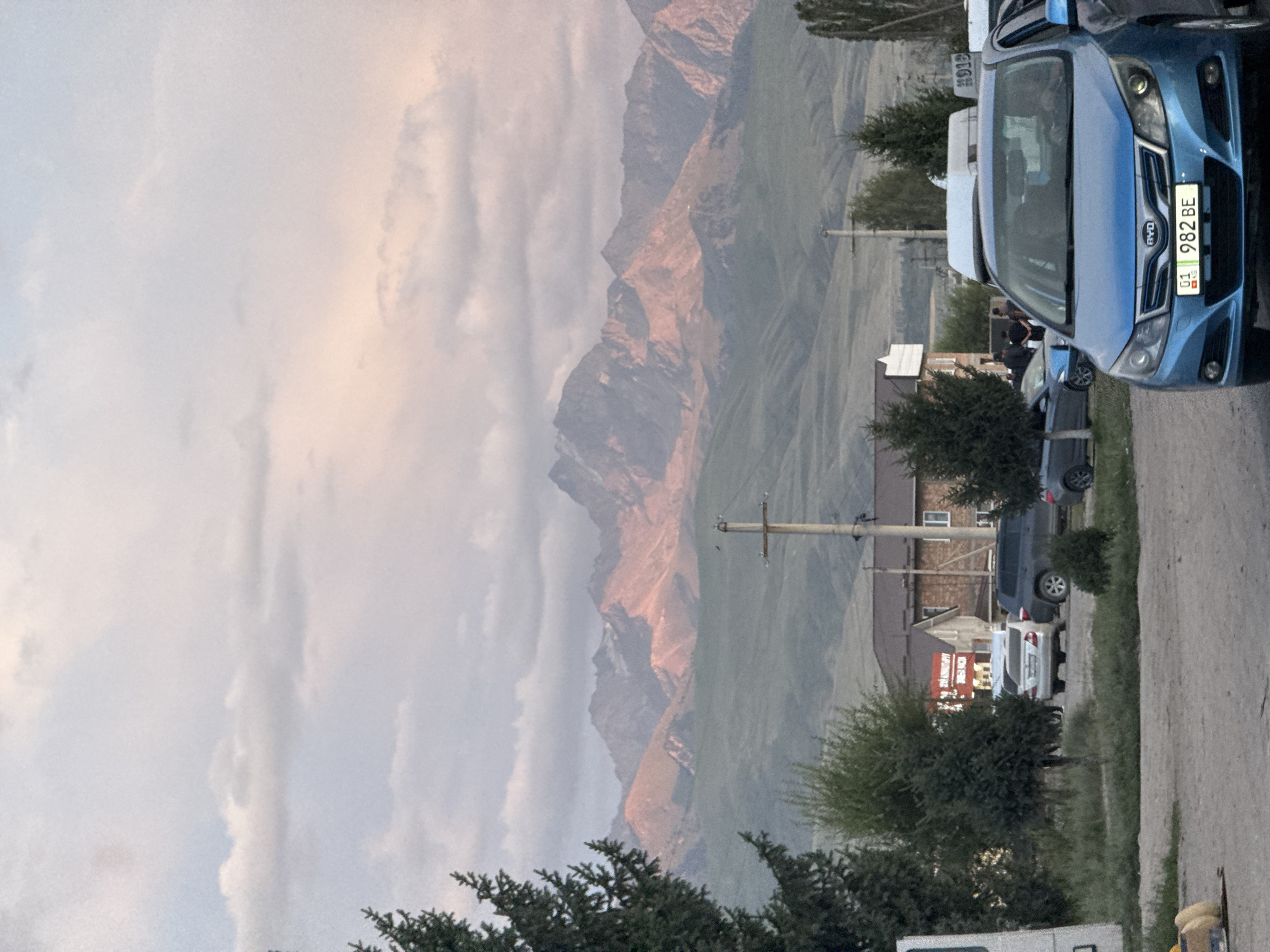 The image depicts a street scene in the city of Zhany-Aryk, Kyrgyzstan. In the foreground, a blue BYD sedan with license plate "01 982 BE" is parked, facing forward. To the left of the sedan, other vehicles and trees are visible. In the middle ground, several buildings are present, including a two-story structure with a dark-colored roof. A power pole with wires stands near the buildings. In the background, a mountain range is prominent, with its peaks illuminated by the setting sun, casting a warm glow on the landscape. The sky above is partly cloudy, with hues of pink and orange from the sunset. Several individuals are standing near the buildings, appearing to be engaged in conversation or activity. The overall scene suggests a typical evening in a semi-urban area bordering a natural landscape.