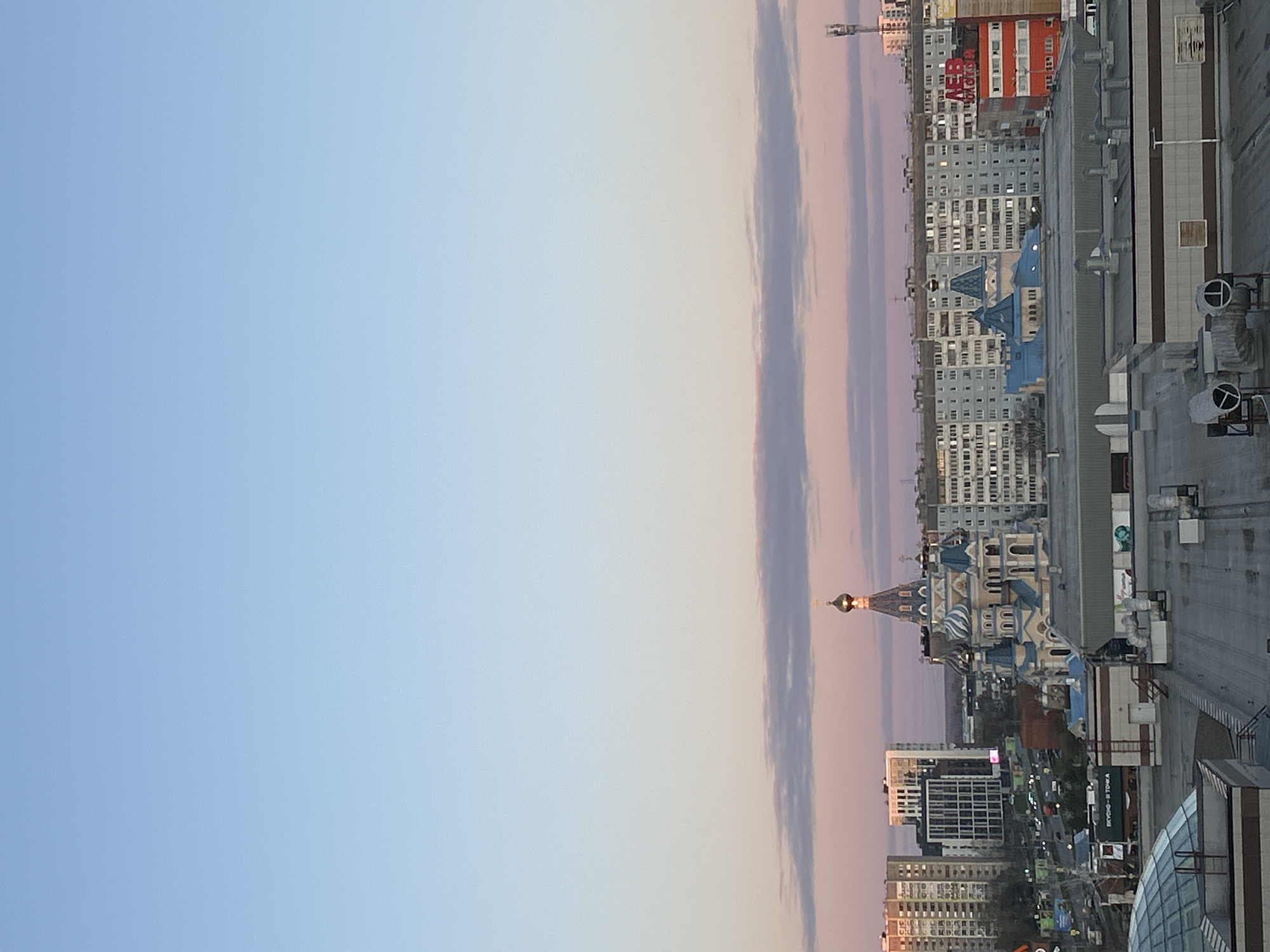 The image shows a daytime view of Izhevsk, Russia, taken from a high vantage point. The sky is predominantly light blue, with pinkish hues near the horizon and wispy clouds. In the foreground, rooftops of buildings are visible. In the distance, multi-story buildings, apartments, and a building with a blue-domed church on top are seen. The light from the sun is reflected on the golden domes. The buildings and streets are in a vertical orientation.