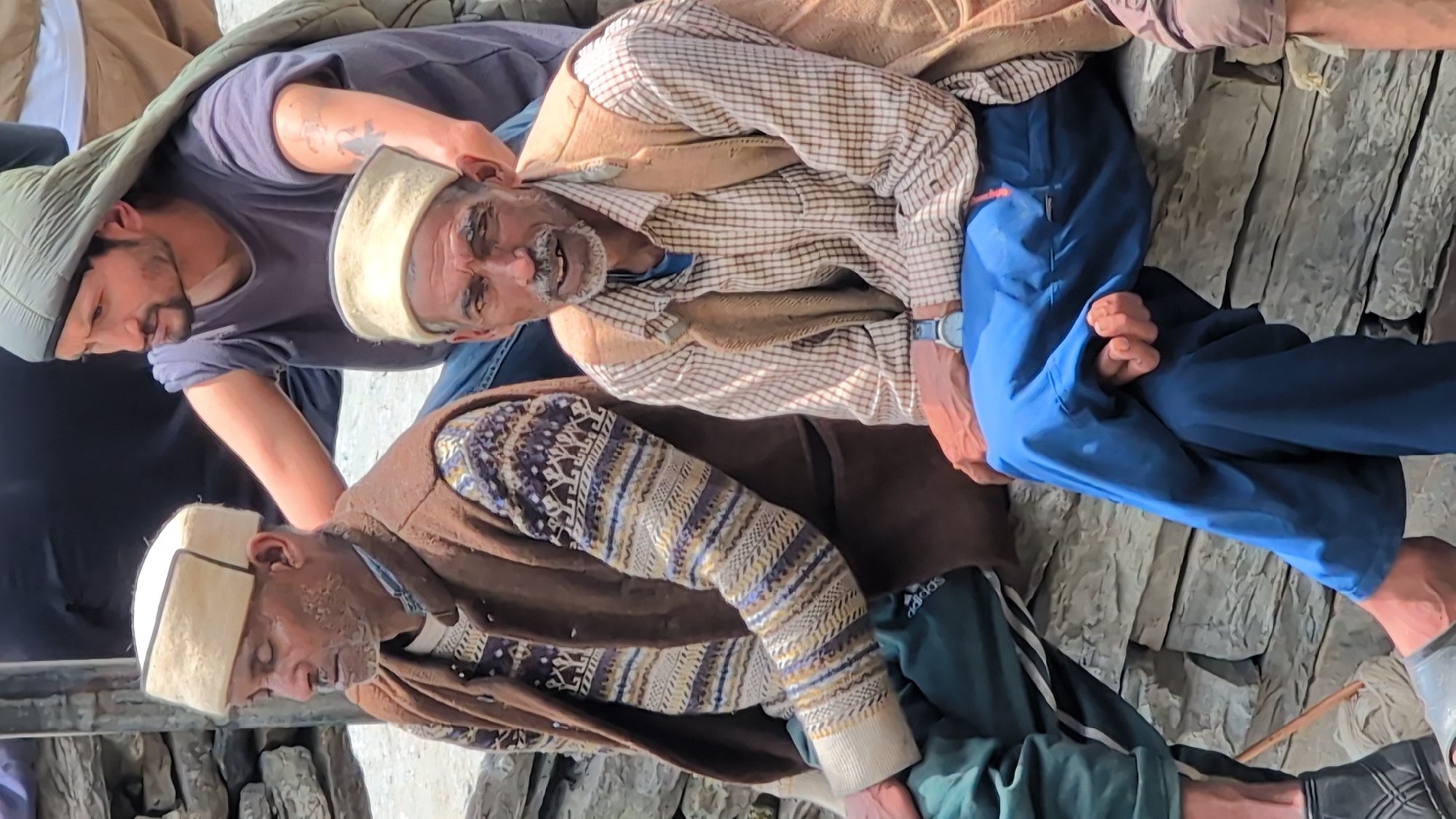 Two men are seated on a stone wall in Malana, India. The man on the left is older, with a light-colored cap and vest over a patterned sweater and green pants. He is looking downwards. The man on the right, also wearing a light-colored cap, is younger with a beard and mustache. He is wearing a plaid shirt, a vest, and blue pants. He is looking towards the viewer with a slight smile. A third person, wearing a grey t-shirt and a green headscarf, is standing behind them, with one arm resting on the shoulder of the man on the left. The background consists of a stone wall and some blurry figures.