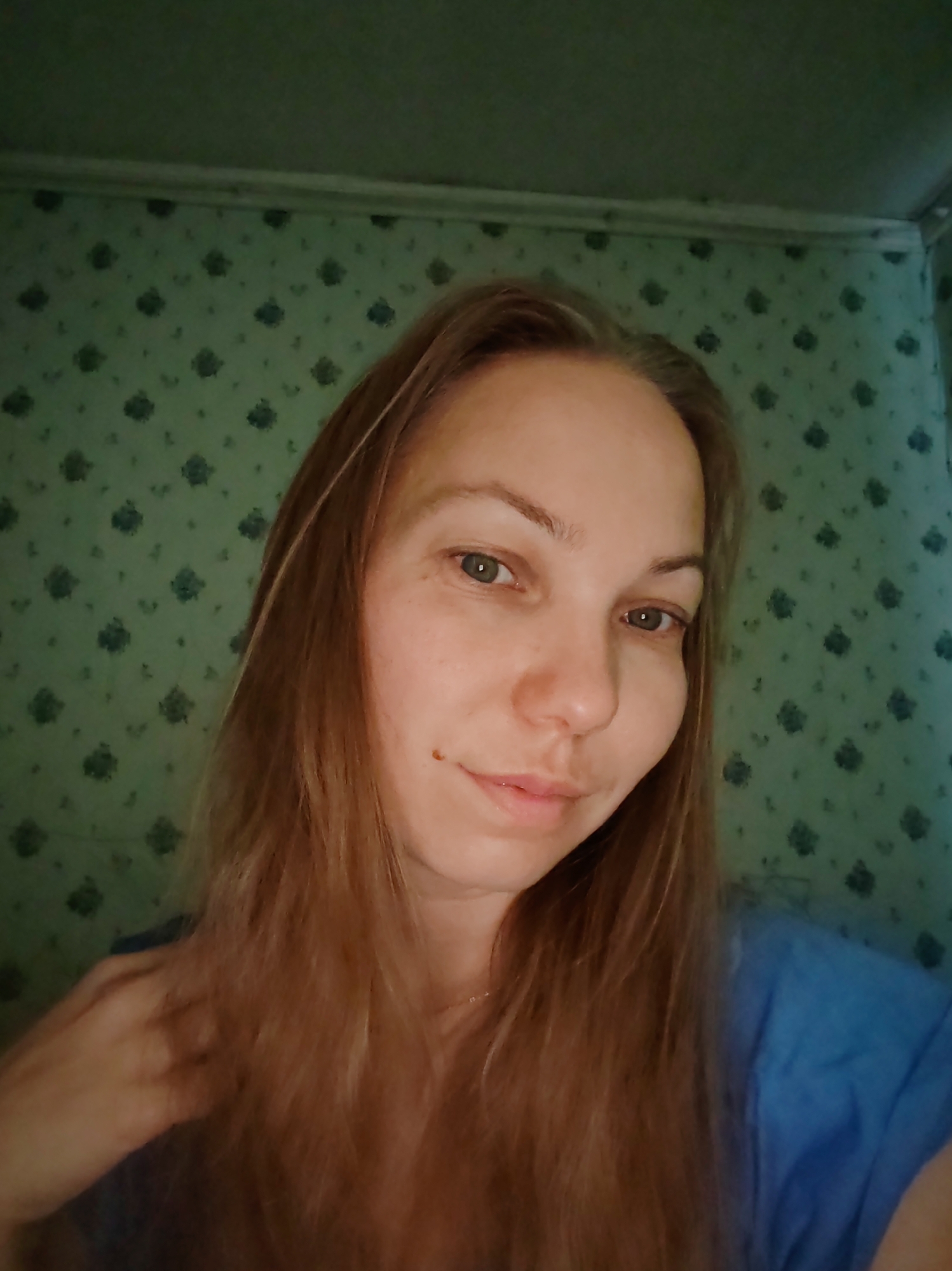 A fair-skinned woman with shoulder-length auburn hair and green eyes is looking directly at the camera. She has a small mole on her left cheek. She is wearing a blue t-shirt. The background is a green patterned wallpaper. The image appears to have been taken indoors, possibly in a residential setting. There is no specific indication of the location within the city of Чухлэм, Russia.