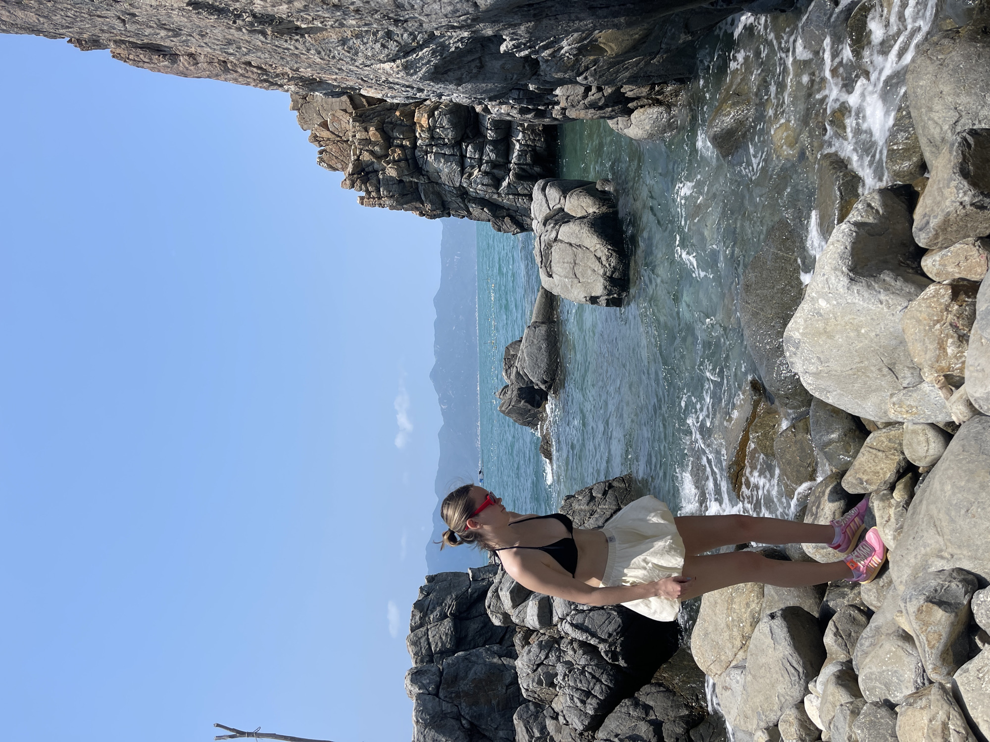 A woman stands on a rocky shore next to clear water in Vĩnh Lương, Vietnam. She wears a black bikini top, a light-colored skirt, and pink sneakers. The shore is composed of large, grey rocks, some wet from the water. A rocky cliff formation rises on the left, and in the distance, mountains are visible. The sky is clear and blue.