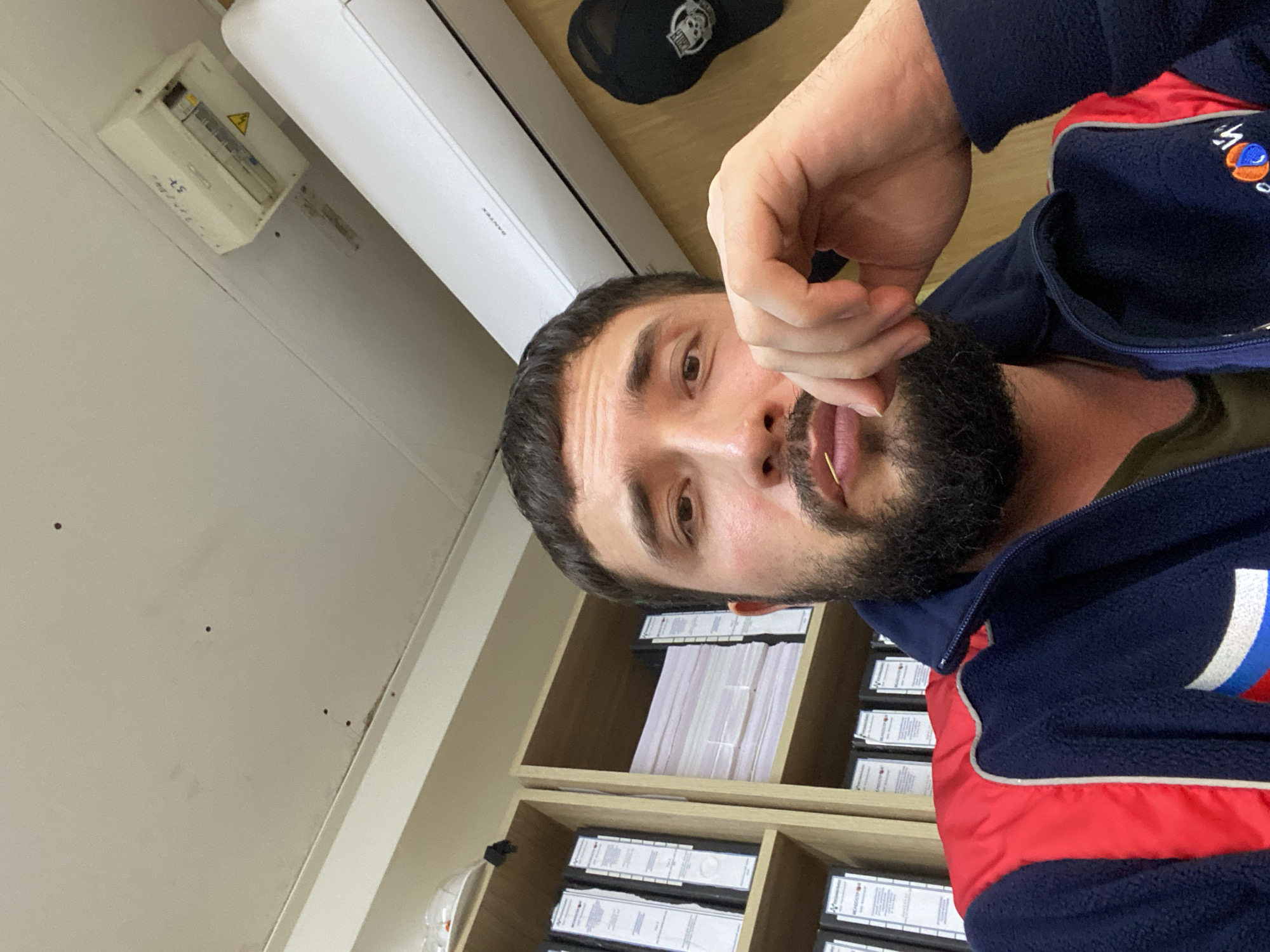 The image shows a man with a beard and mustache, wearing a dark blue and red jacket, sitting in an office. He is holding a toothpick to his lips. Behind him, shelves are filled with binders and papers. An air conditioner is mounted on the wall above him, and a black baseball cap with a skull logo hangs on the wall to the right. The office appears to be in Luzhitsy, Russia, as indicated by the text on the air conditioner unit. The lighting suggests an indoor setting, likely during the day.