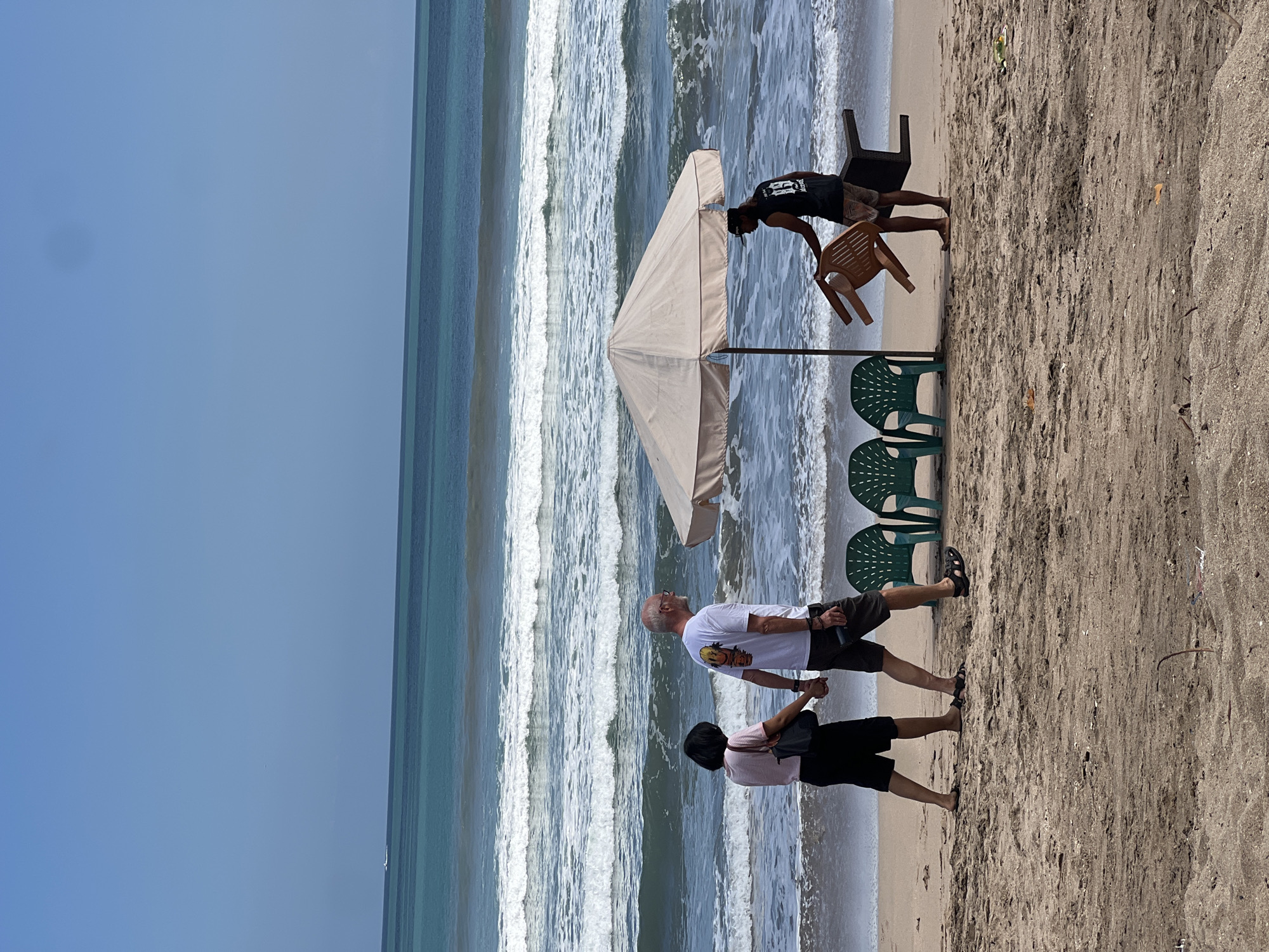 In Kuta, Bali, Indonesia, a couple walks hand-in-hand on a sandy beach, their backs to the viewer. The woman, on the left, wears a pink and white striped top and black shorts, carrying a black purse. The man, to her right, is in a white t-shirt and dark shorts, holding a mobile phone.
Further along the beach, under a light-colored umbrella, a person wearing a black t-shirt and patterned shorts is stacking chairs. Two green plastic chairs are already placed on the sand to the left of the umbrella, and another brown plastic chair is being held by the person in black. The ocean behind them has moderate waves and a clear blue sky above.