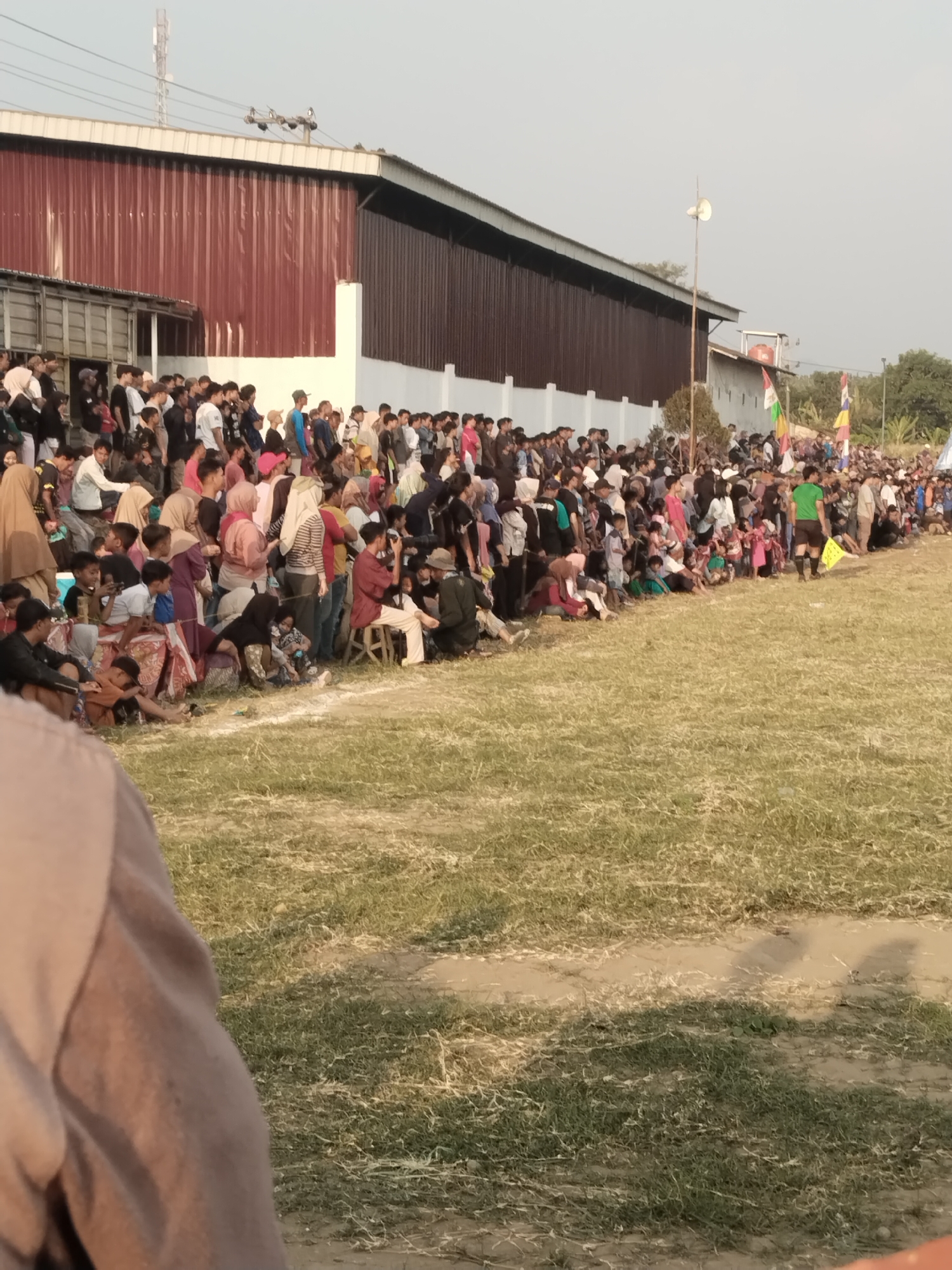 The image captures a crowd of people gathered outdoors in Margasari, Indonesia, likely for an event or sporting match given the presence of flags and what appears to be a field. A large, dark red building with a white trim runs along the left side of the frame, possibly a factory or warehouse. In front of the building, a diverse group of individuals, including men, women, and children, are seated and standing. Many are dressed in casual attire, with some wearing headscarves. The ground in front of the crowd is covered in dry grass with some patches of bare earth. In the background, there are trees and other structures, including a tall antenna. The sky is a light, hazy blue, suggesting it might be late afternoon or early morning. The overall scene depicts a public gathering in a semi-urban or industrial area.