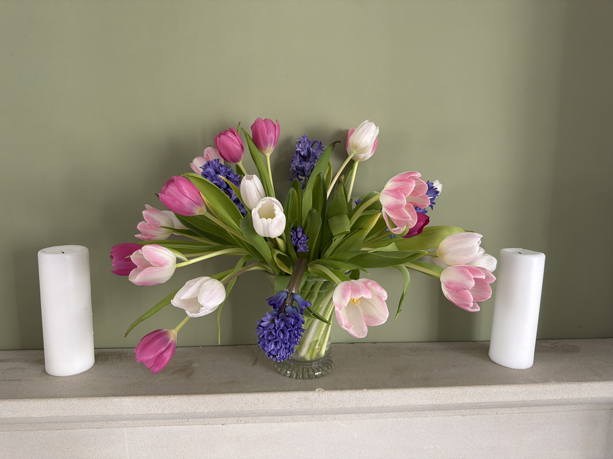 A still-life scene shows a bouquet of tulips and hyacinths in a glass vase, flanked by two white pillar candles. The flowers are in shades of pink, white, and purple, with green stems and leaves. The arrangement sits on a gray shelf, which is presumably a mantelpiece due to its placement against a muted green wall. The setting appears to be indoors, and while the specific location in Malvern Hills, United Kingdom, is not discernible from the image, it is likely a residential interior given the decor. There are no people or actions evident in the photograph, which focuses on the arrangement of the flowers and candles as its subject.