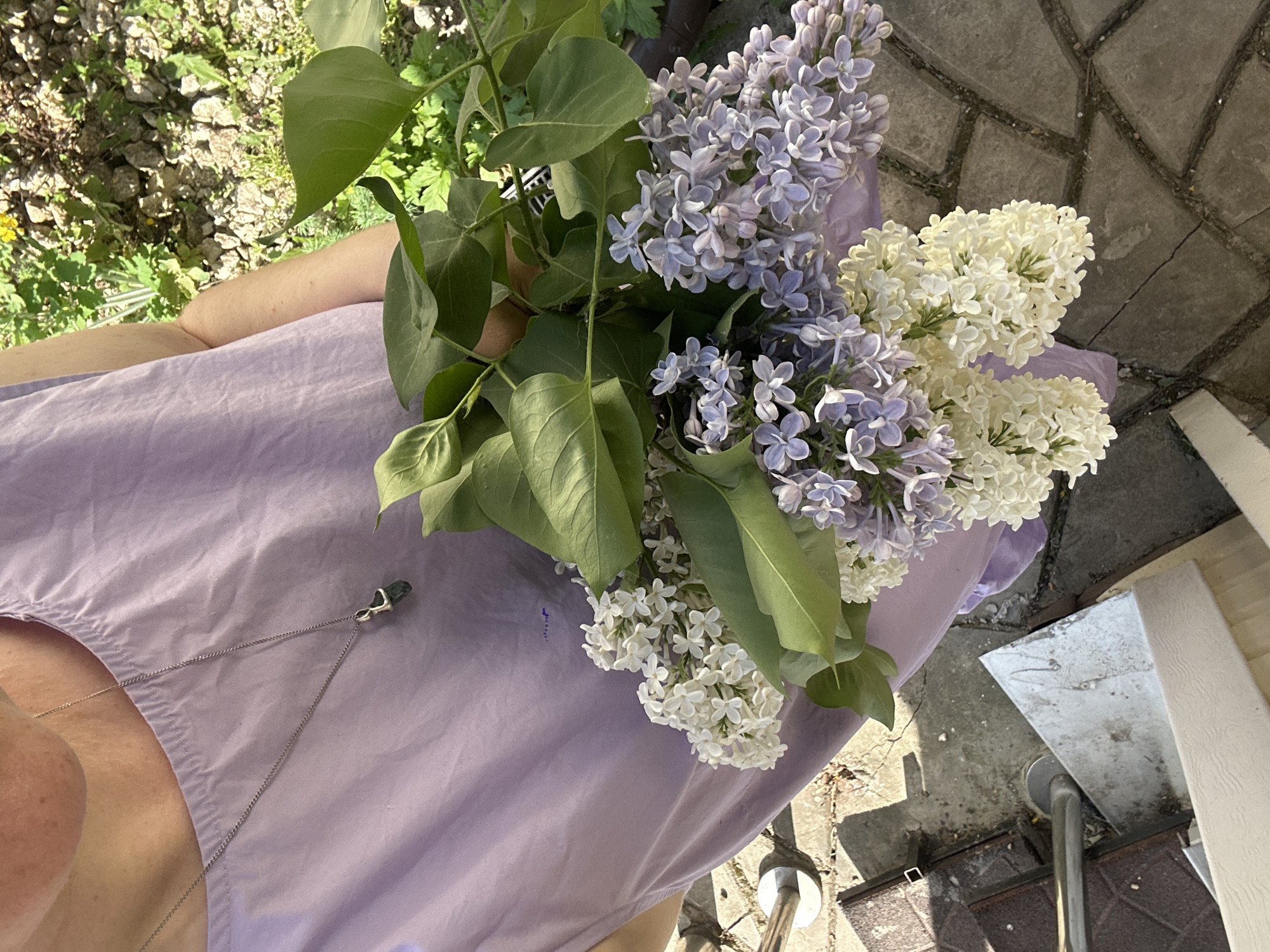 A person wearing a lavender dress and a silver necklace with a dark green pendant holds a bouquet of lilacs. The bouquet contains both light purple and white lilacs with green leaves. The person is standing on a paved surface, possibly a sidewalk or patio, with a view of greenery (grass and plants) and possibly a brick or stone wall in the background. The person's face is partially visible. The location is unknown and cannot be determined from the image.
