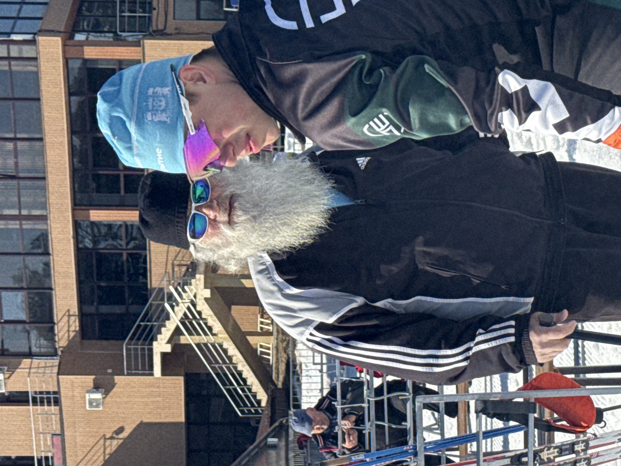 Two men stand in the foreground. The older man has a long white beard and wears sunglasses, a black hat, and an Adidas jacket. The younger man wears a blue beanie and colorful sunglasses. Both appear to be on a ski lift. An orange bag is on the ski lift in front of the older man. In the background is a multi-story brick building with windows and a metal staircase. Another person is visible in the background. The location is possibly Прикамский, Russia.
