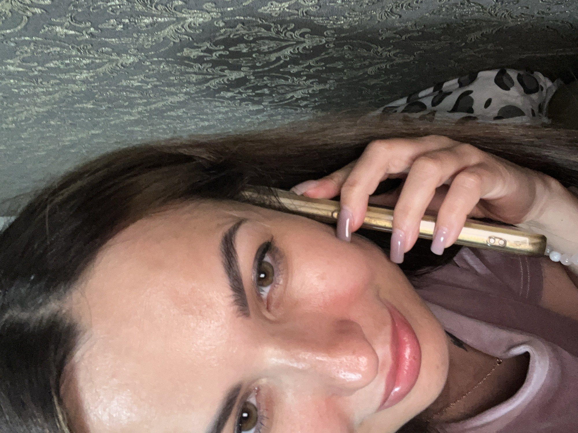 A close-up selfie of a woman holding a gold-colored phone to her ear, taken indoors against a grey, patterned wallpaper. The woman has brown hair and is wearing a pink t-shirt. Her nails are painted a light purple color. The lighting in the image is warm and casts highlights on her face.