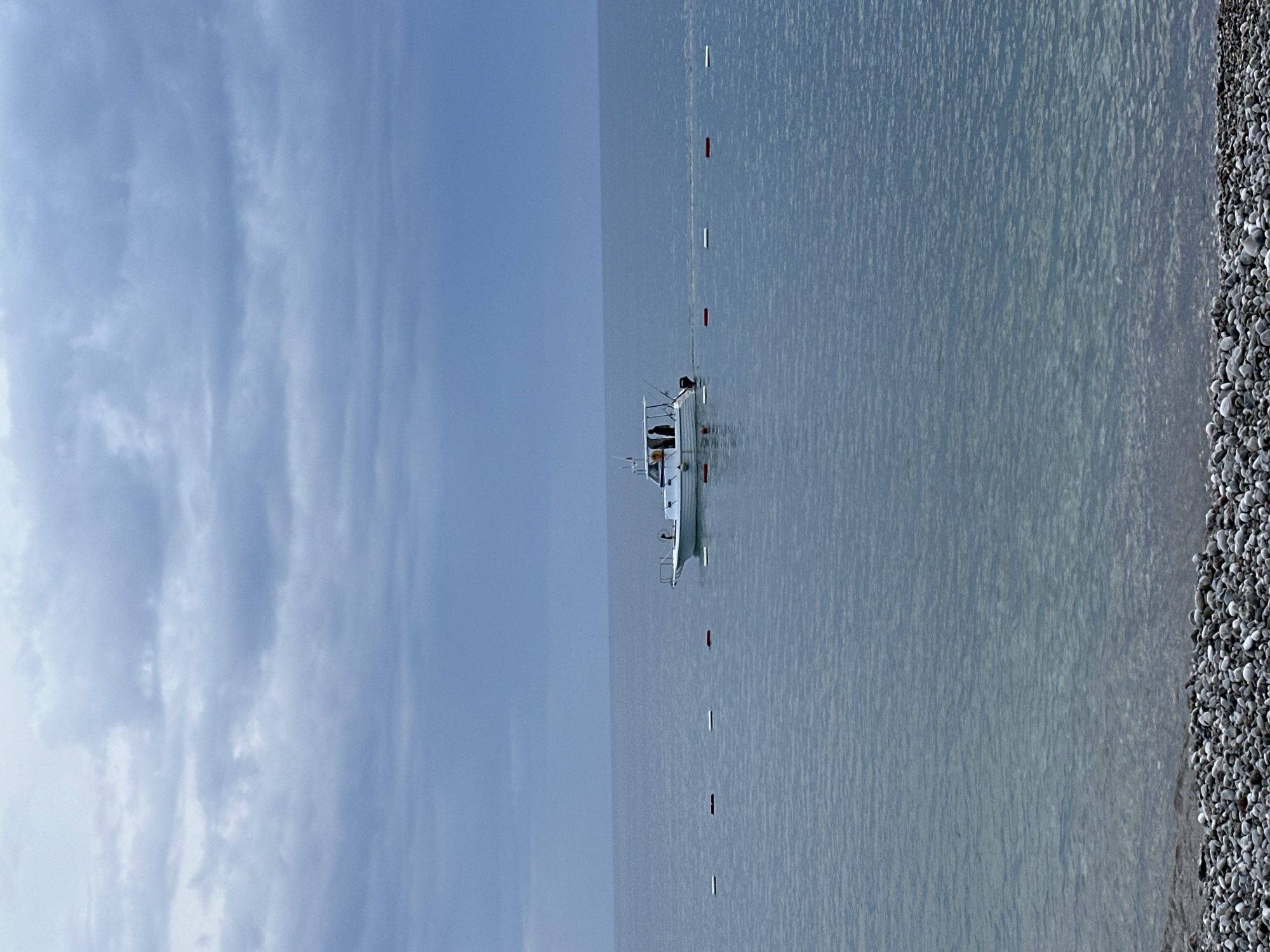 The scene shows a calm sea off the coast of Šušanj, Montenegro, under a cloudy sky. A small white boat is anchored in the water, marked by a line of red and white buoys extending towards the shore. A pebbly beach borders the water on the right side of the image. No people are visible in the shot.