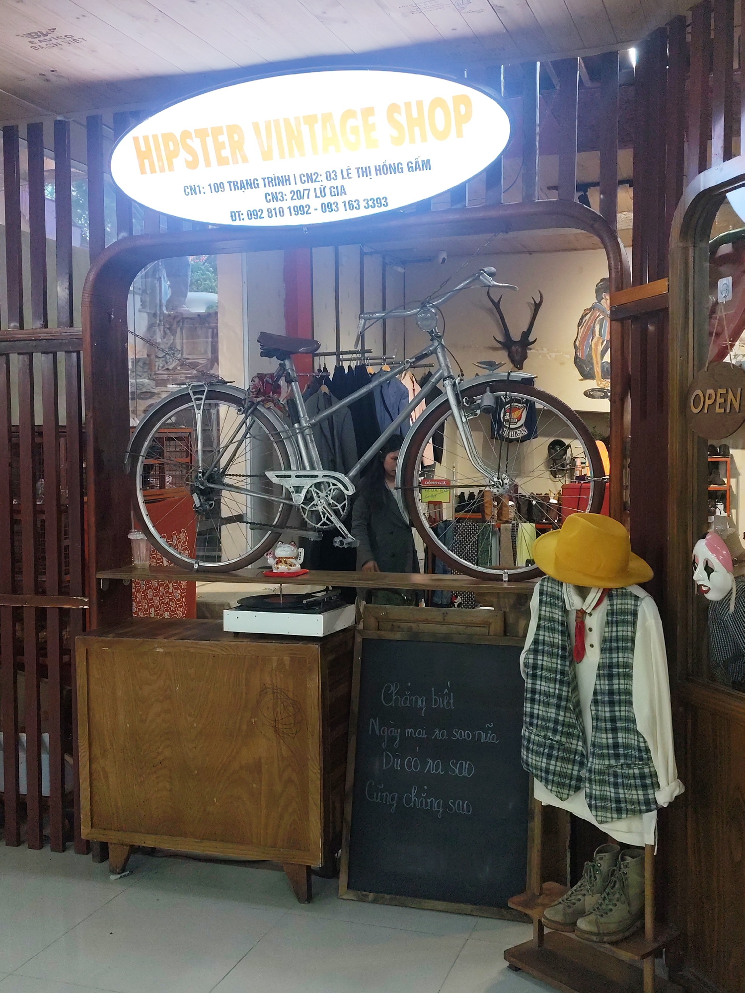The image depicts the exterior of "Hipster Vintage Shop" in Da Lat, Vietnam. A large oval sign with yellow lettering, reading "HIPSTER VINTAGE SHOP" and store contact information, is prominently displayed above the entrance. The shop features a rustic, wooden facade with vertical planks. Inside, a vintage bicycle is displayed, suspended from the ceiling or a rack. Clothing items are hung on a rack behind the bicycle. A mannequin wearing a plaid vest, a white shirt, and a red ascot is positioned in the foreground, topped with a yellow fedora. A pair of tan combat boots are displayed on a wooden stand beneath the mannequin. To the left of the mannequin, a blackboard with handwritten Vietnamese text is visible. The background reveals more of the shop's interior, including shelves with various items and a mounted deer head. The floor is tiled with a light-colored pattern. The overall scene suggests a vintage-themed retail establishment.