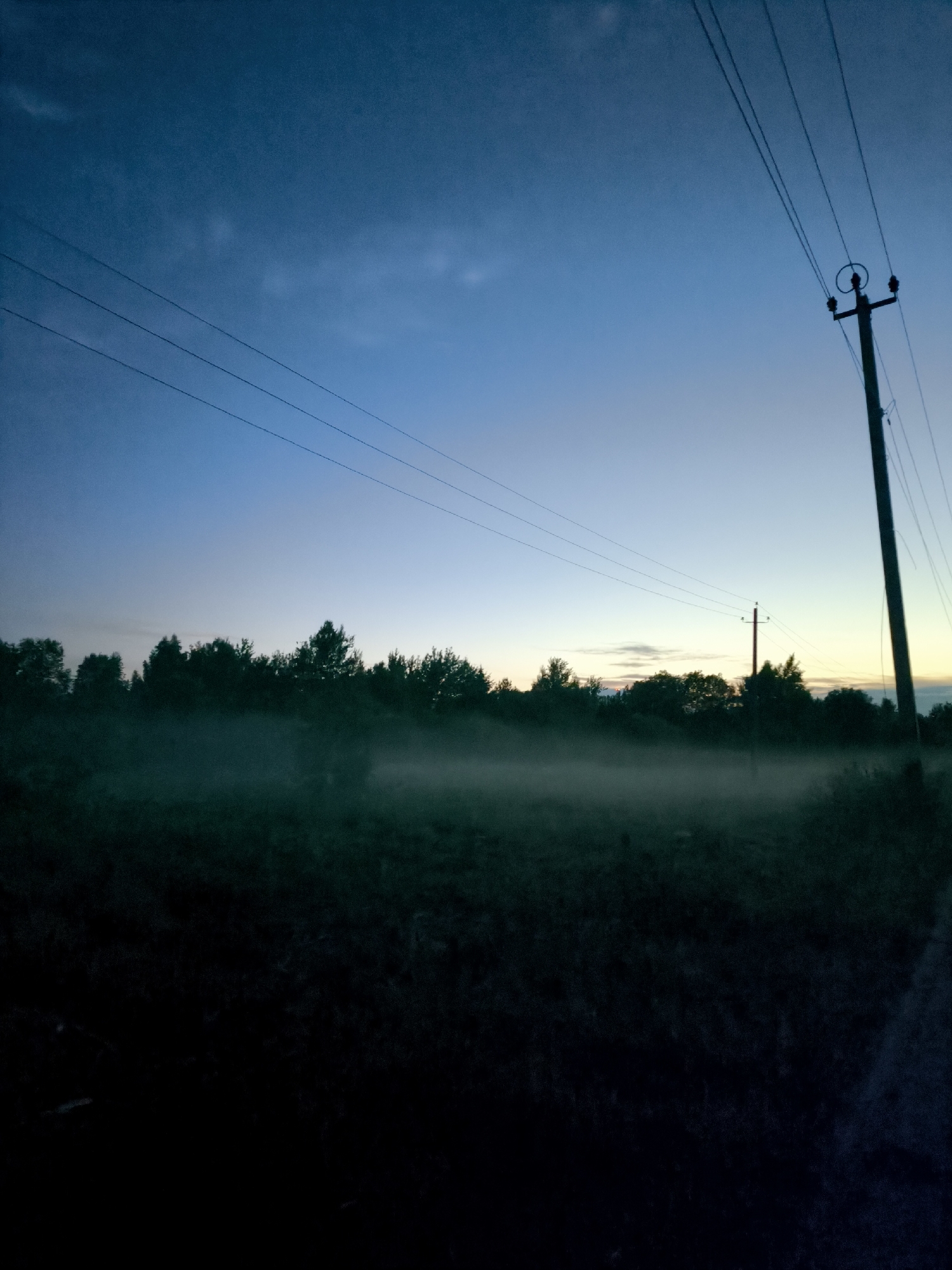 The image depicts a rural landscape at dusk. A telephone pole stands tall on the right side of the frame, with wires extending diagonally across the dark blue sky. Below, a line of silhouetted trees forms a dark horizon. A wispy layer of fog or mist hovers low over a field in the foreground, obscuring the ground and creating a soft, ethereal effect. The overall atmosphere is serene and quiet, with the fading light casting long shadows and a sense of stillness.