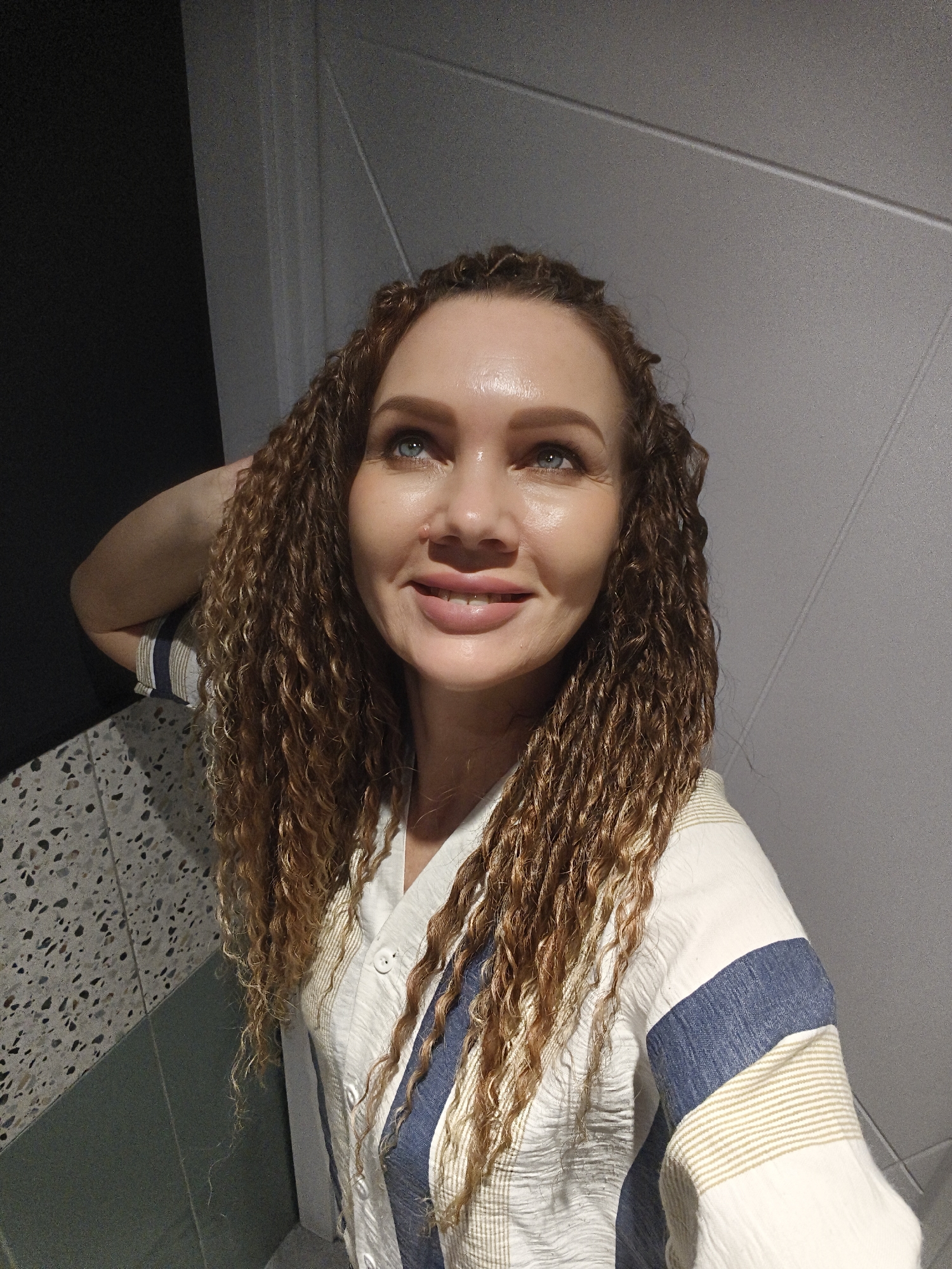 A light-skinned woman with light blue eyes and curly auburn hair is smiling and looking up. She is wearing a white, button-down shirt with blue and beige stripes on the sleeves and front. She has her left arm raised and her hand resting on the back of her head. She is standing next to a countertop with a gray and white speckled surface, and a wall with light gray panels. To the left, there is a dark, undefined area. The location is Unknown, Unknown.