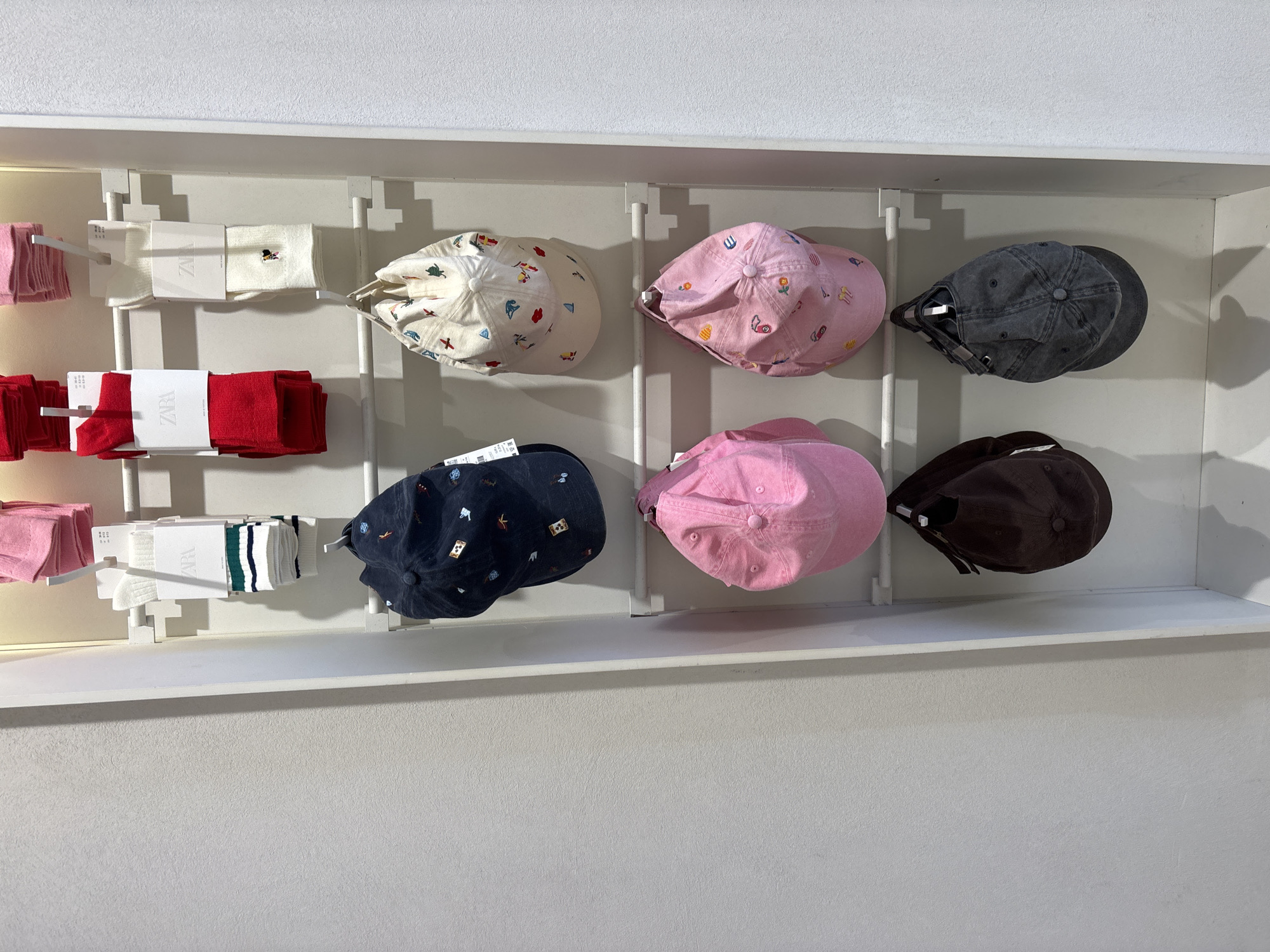 A clothing display is visible in an indoor setting. Racks affixed to the wall showcase assorted merchandise. The left side displays folded clothing or fabric items in pink and red colors alongside wrapped sock sets. To the right, a selection of baseball caps is arranged in two rows, offering a variety of colors including white with prints, pink, blue with prints, gray, and brown. The backdrop is a plain, light-colored wall, and the overall impression is of a retail environment. There is no evidence of location in Sofia, Bulgaria.