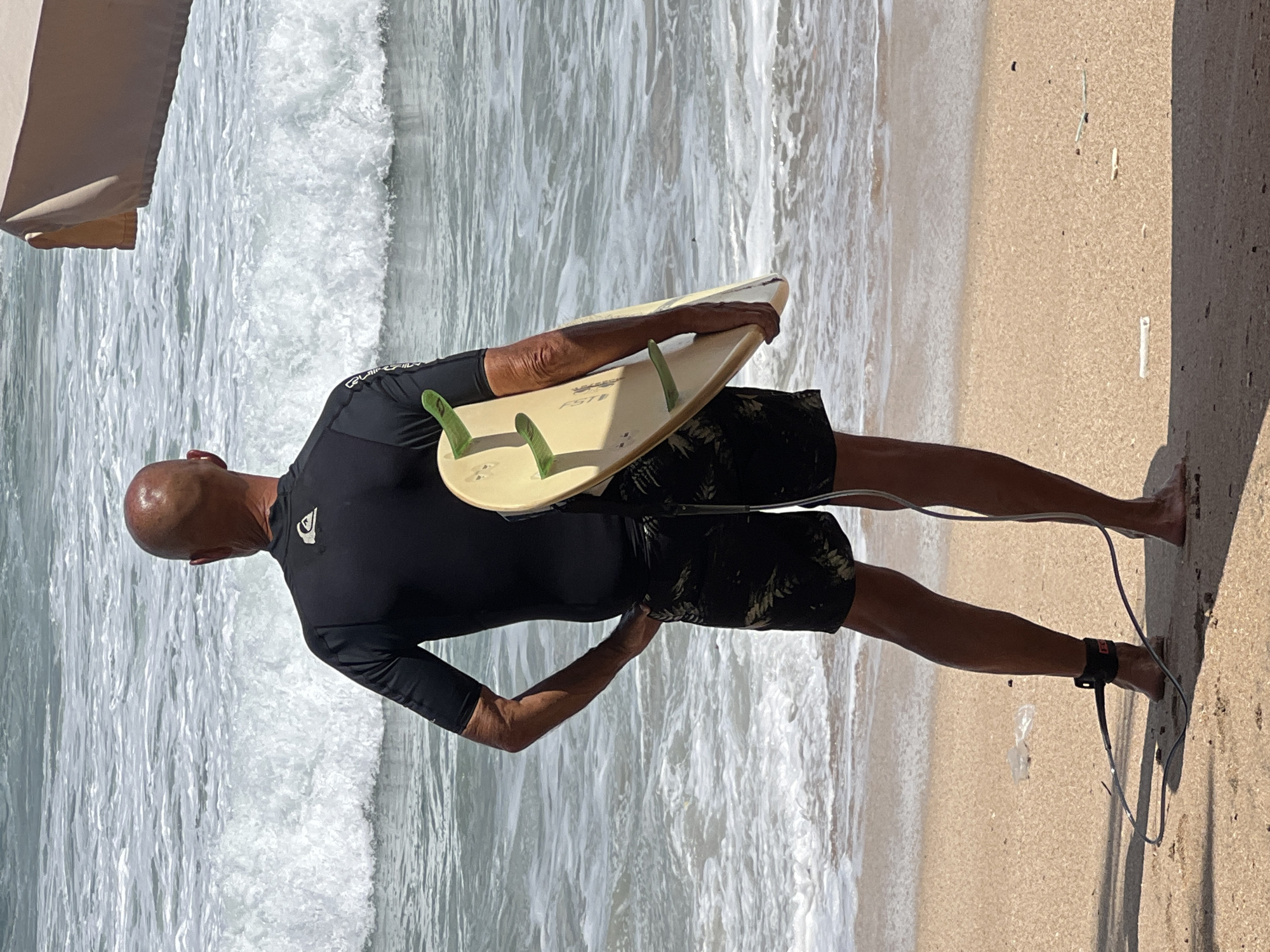 A bald, fair-skinned man stands on a sandy beach, facing away from the camera and towards the ocean. He is wearing a black wetsuit top and black patterned swim trunks. The man holds a pale yellow surfboard vertically under his right arm. The surfboard has three green fins visible. His left hand is in his swim trunk pocket. A black leash connects from the surfboard to his left ankle, where it is secured by a black ankle strap. The ocean is a light blue-gray with white waves breaking near the shore. A light brown beach umbrella is partially visible at the top right of the frame. The sand is light brown with some small debris. The scene appears to be located in Kuta, Indonesia.