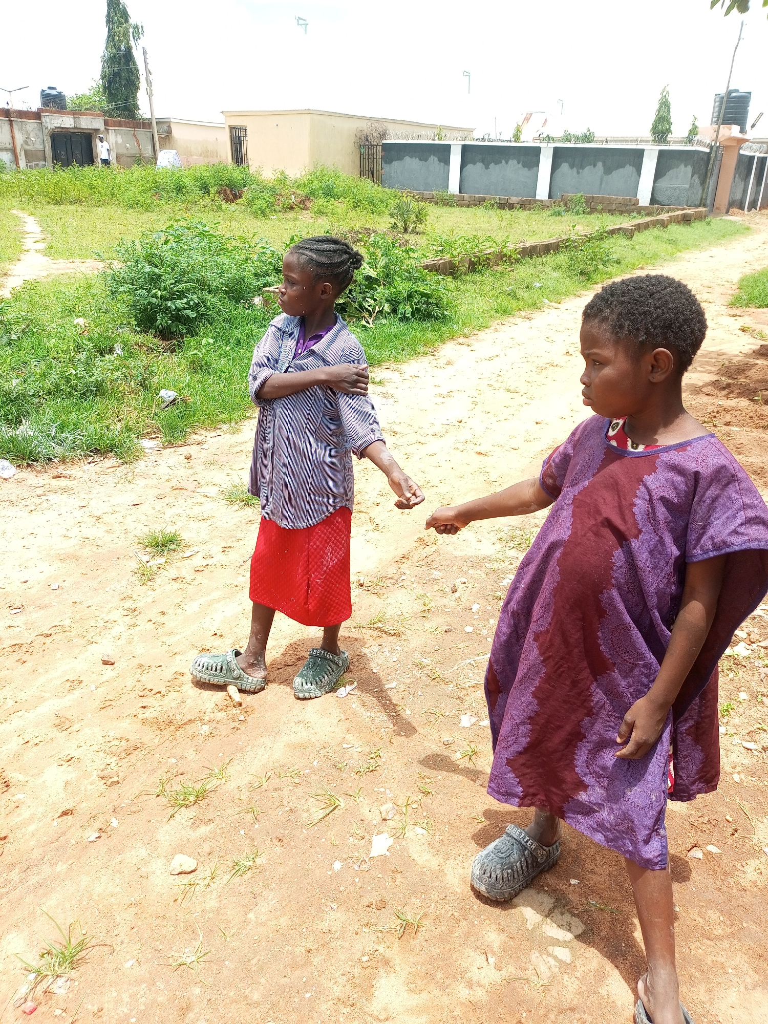 In an outdoor scene in Tempure, Nigeria, two young girls stand on a dirt path. The girl on the left, facing away from the camera, wears a striped blue and white shirt over a purple undershirt and a red skirt. She has braided hair. The girl on the right, facing left, is wearing a purple garment with a patterned design. Both girls are wearing grey clogs. The background shows a dirt road, green vegetation, and several low-rise buildings. The sky is overcast.