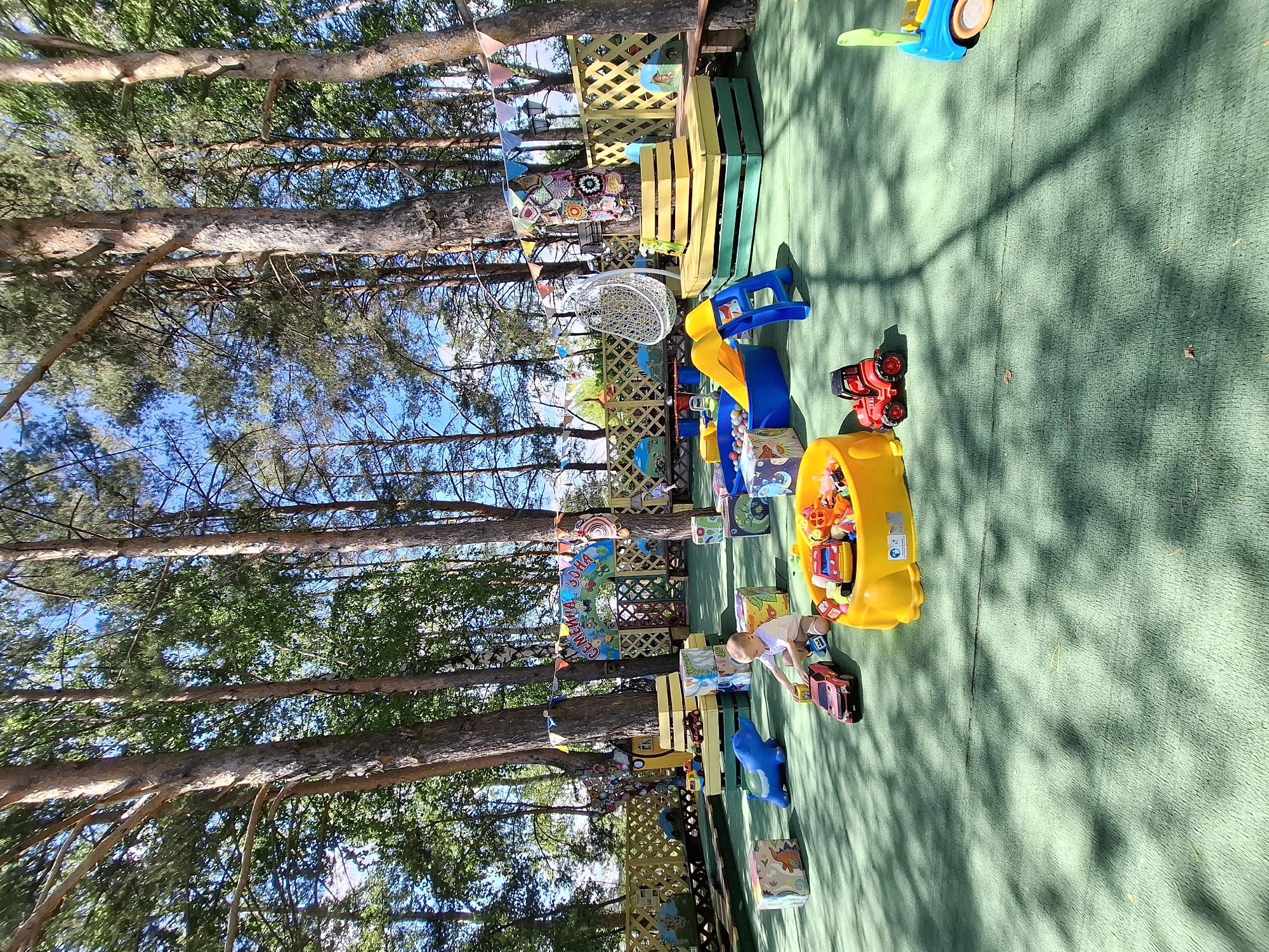 In a children's play area in Chaikovsky, Russia, a young boy sits on a green artificial turf surface, playing with toy cars. He is wearing a white shirt and beige shorts. In front of him, a yellow sandbox is filled with various toys, including cars and blocks. Nearby, a red toy tractor and a red toy car are also present. To the left, a blue elephant-shaped toy is on the ground, and several colorful blocks are scattered around the play area. Behind the boy, there is a blue plastic slide and a ball pit. The play area is set within a grove of tall pine trees, with a wooden fence and decorative banners visible in the background. Sunlight filters through the trees, casting shadows on the ground. The overall impression is of an outdoor recreational space designed for children.