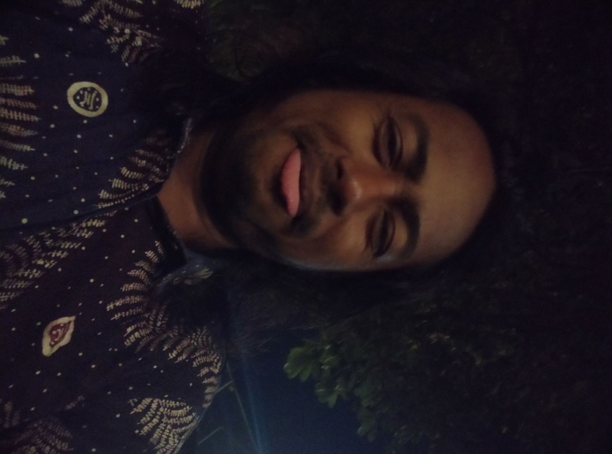 The image shows a close-up, selfie-style shot of a person with dark skin and long, dark hair, wearing a dark blue patterned shirt. The person's face is partially obscured by shadows and they are sticking out their tongue. The background appears to be outdoors at night, with dark foliage visible. The shirt has a repeating pattern of light-colored, stylized fern-like designs and small white dots. There is a circular pin with a design on the left side of the chest and a leaf-shaped patch on the right side of the chest. The image is taken in a dark environment.