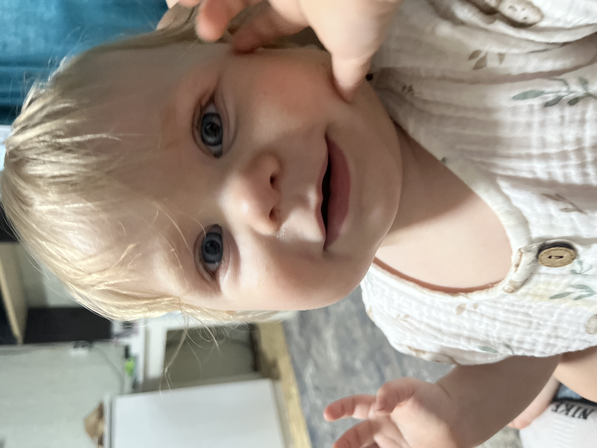 A close-up, head-and-shoulders shot shows a fair-skinned toddler with blonde hair and blue eyes. The child is looking directly at the camera with a slight smile. The child's right hand is raised, and a thumb and index finger are touching the child's cheek. The child is wearing a light-colored, textured shirt with a botanical print. The background is blurred, showing a white appliance, possibly a refrigerator, to the left, and a dark curtain to the right. The setting appears to be indoors, likely a residential interior. The image does not contain any information about the city of Фоки, Russia.