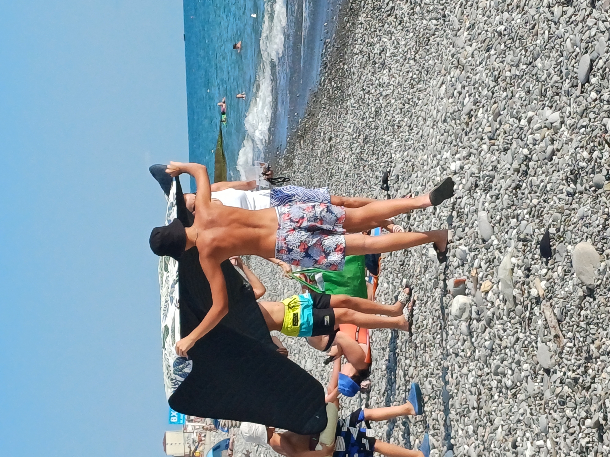 The image depicts a beach scene on a sunny day, likely in Sochi, Russia. Several people, predominantly young men and boys, are visible on a pebble beach. The sea, with gentle waves, is in the background, and the sky is clear blue.
The main subjects are a group of individuals lounging or posing on the beach. One young man in the foreground is lying on his back, partially covered by a black blanket or towel. He is wearing a black cap and patterned swim shorts. Other individuals are also in swim attire, with some lying down and others standing or in motion. One person is wearing bright yellow and blue swim shorts, while another has on patterned blue and white shorts.
The activity appears to be a leisurely day at the beach, with people relaxing, sunbathing, and possibly preparing to go into the water. The presence of a surfboard or bodyboard suggests water sports. The beach is covered in a multitude of grey and white pebbles.
Notable details include the bright sunlight, indicating a clear and warm day. The vibrant blue of the sea and sky are prominent. In the background, a few other people can be seen in the water. The overall mood seems relaxed and summery.
There is some visible text on a blue sign at the bottom of the image, but it is not clear enough to read. The setting is unmistakably a beach, characterized by the pebbles, the sea, and the people dressed for swimming and sunbathing.