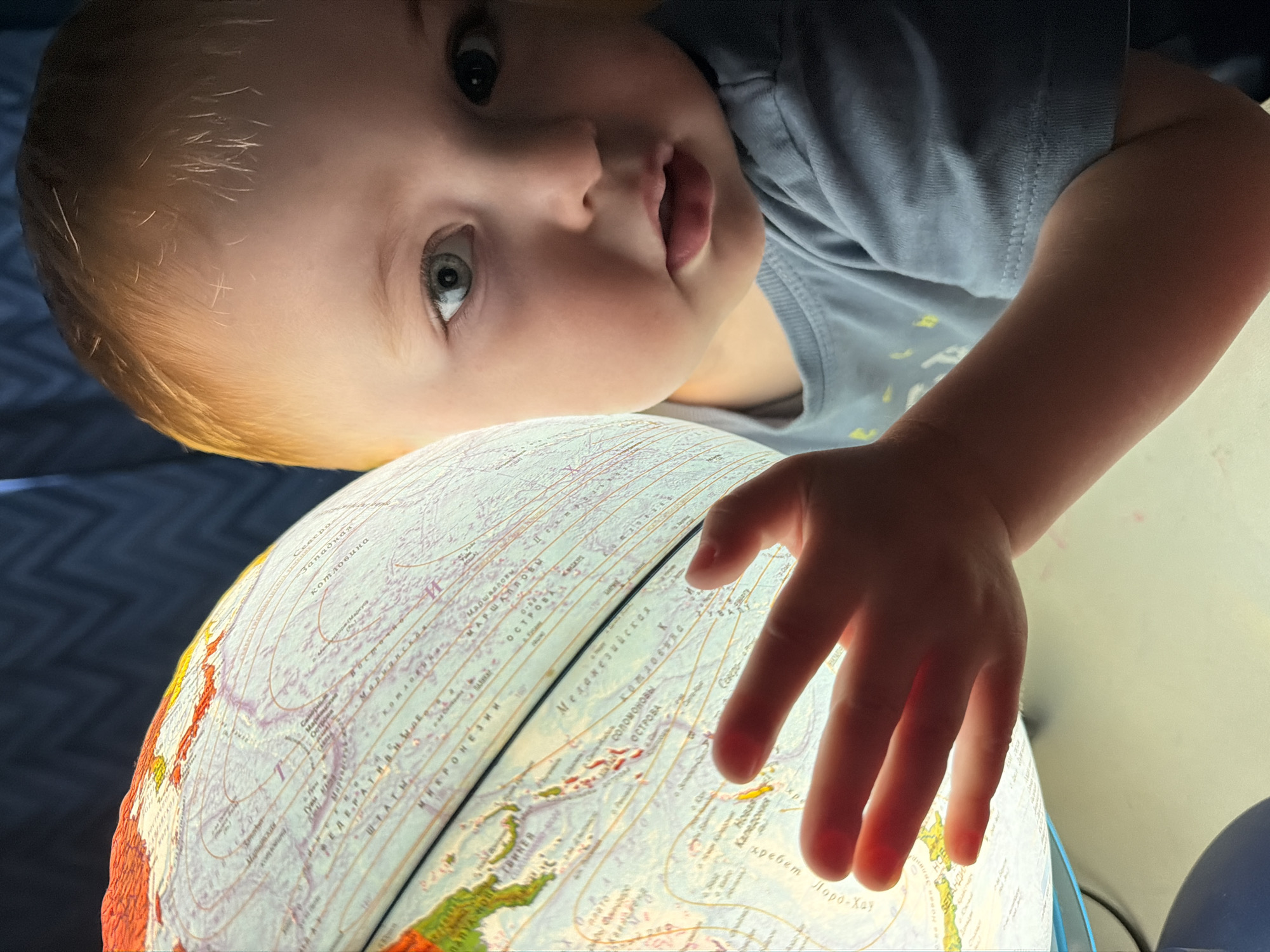 In an indoor setting, a young child with fair skin and red hair is shown with a portion of their face and hand visible in the foreground. The child is looking towards the viewer with wide, light-colored eyes. Their right hand is extended and touching a backlit globe, which displays geographical markings and text in Russian. The globe is illuminated from within, casting a warm glow. The background consists of a dark, textured surface, possibly a curtain or wall. The environment suggests a home or educational space. The image does not contain any specific information that would allow for the determination of a location in Votkinsk, Russia.