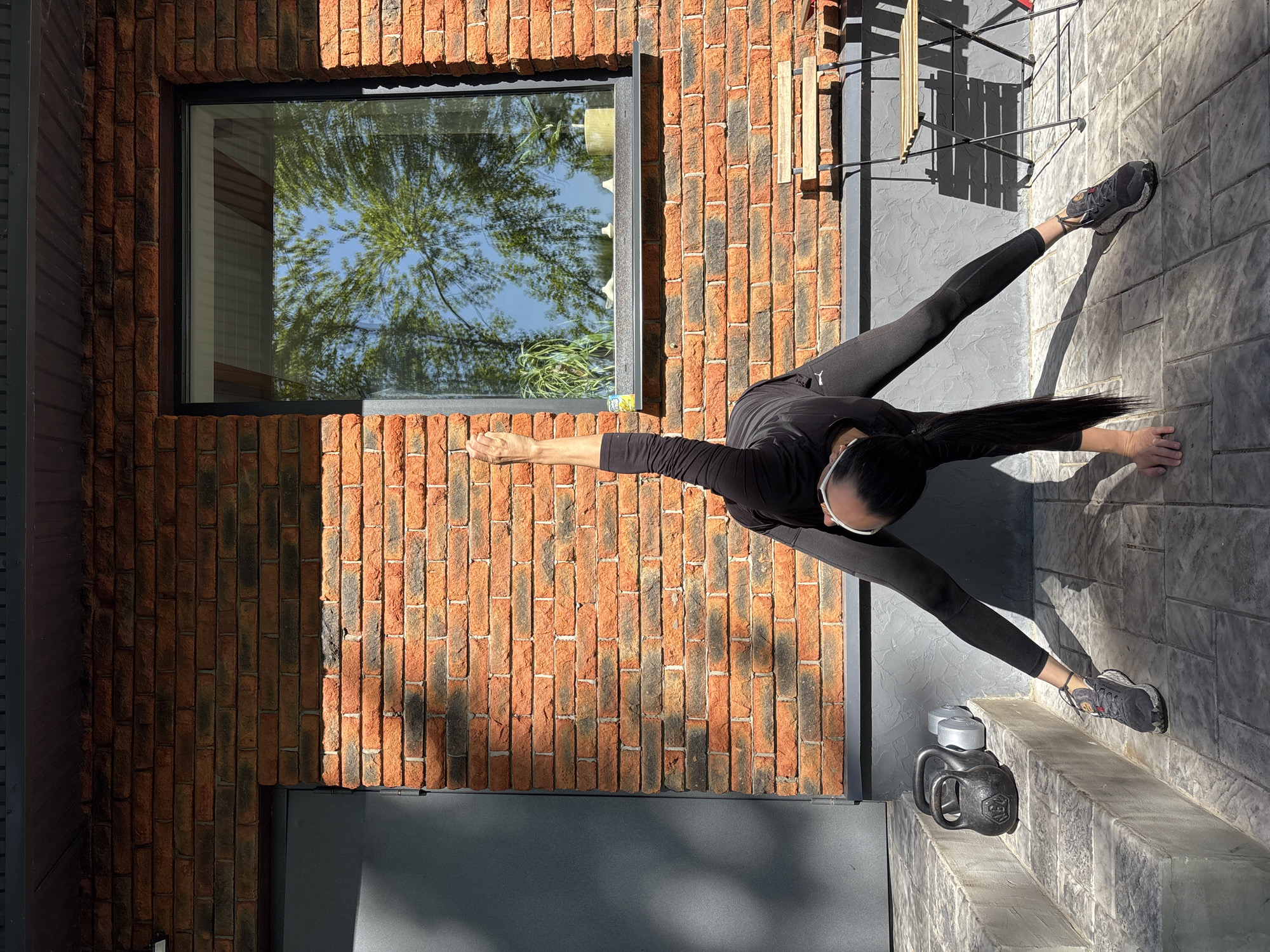 A person is stretching outdoors on a concrete surface adjacent to a brick building. The person is wearing black athletic clothing and sneakers, with their legs spread apart in a wide stance and one arm extended towards their foot. A window reflects a tree and blue sky. A kettlebell and weights are on the surface near the person's foot. A wooden chair is partially visible in the background. The location is Chaikovsky, Russia.