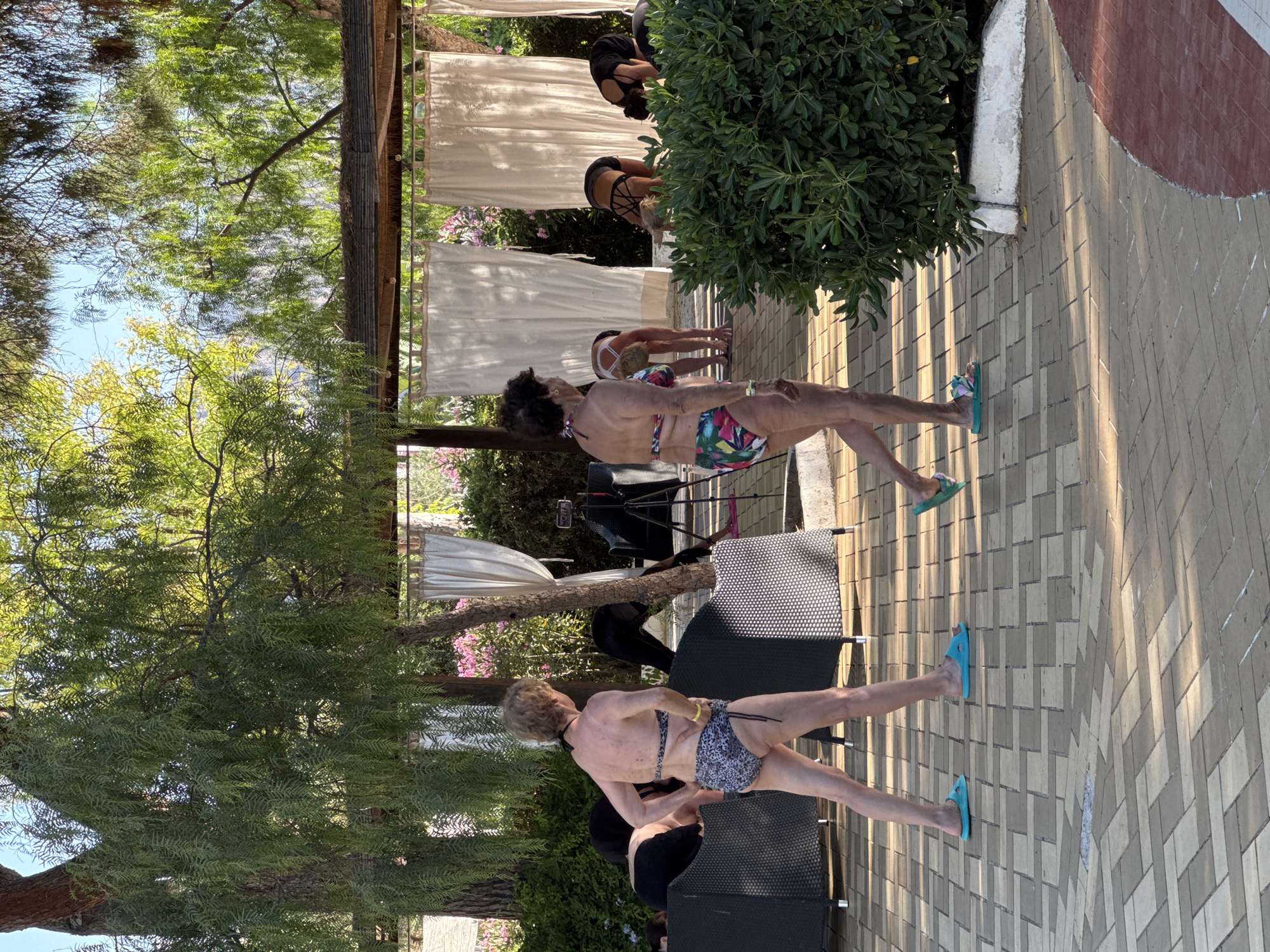 In Isola delle Femmine, Italy, a group of women are engaged in a yoga session outdoors. The scene is set on a paved courtyard, possibly at a resort or public space, with lush green foliage and trees providing a natural backdrop. In the foreground, two women are walking away from the camera, one wearing a leopard-print bikini and the other a floral-patterned bikini. Several other women are visible in the mid-ground, in various yoga poses, facing away from the camera, with white curtains forming a backdrop behind them. A tripod with a phone is set up, suggesting the session might be recorded or live-streamed. The overall atmosphere suggests a serene and active outdoor activity.