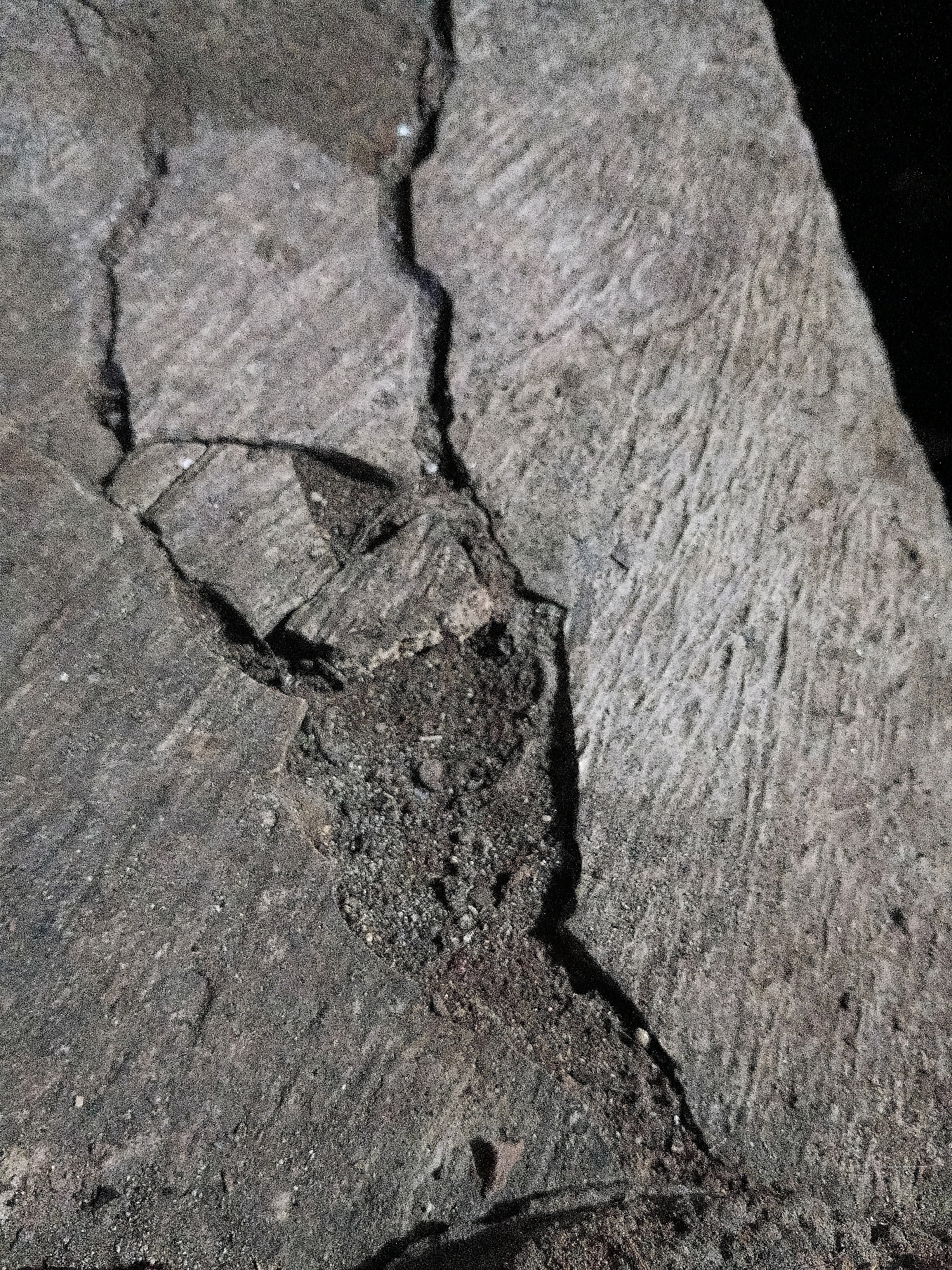 The image displays a close-up, overhead view of a cracked stone surface. The stone is rough and textured, with distinct parallel striations indicating it may have been carved or worn. A dark, irregular crack runs diagonally across the surface, filled with dark debris. Another smaller crack branches off from the main one. The lighting suggests it is outdoors, with shadows cast on the stone, but no specific location within the city of Cikal, Indonesia can be determined from the image itself. There are no people or other living subjects visible.