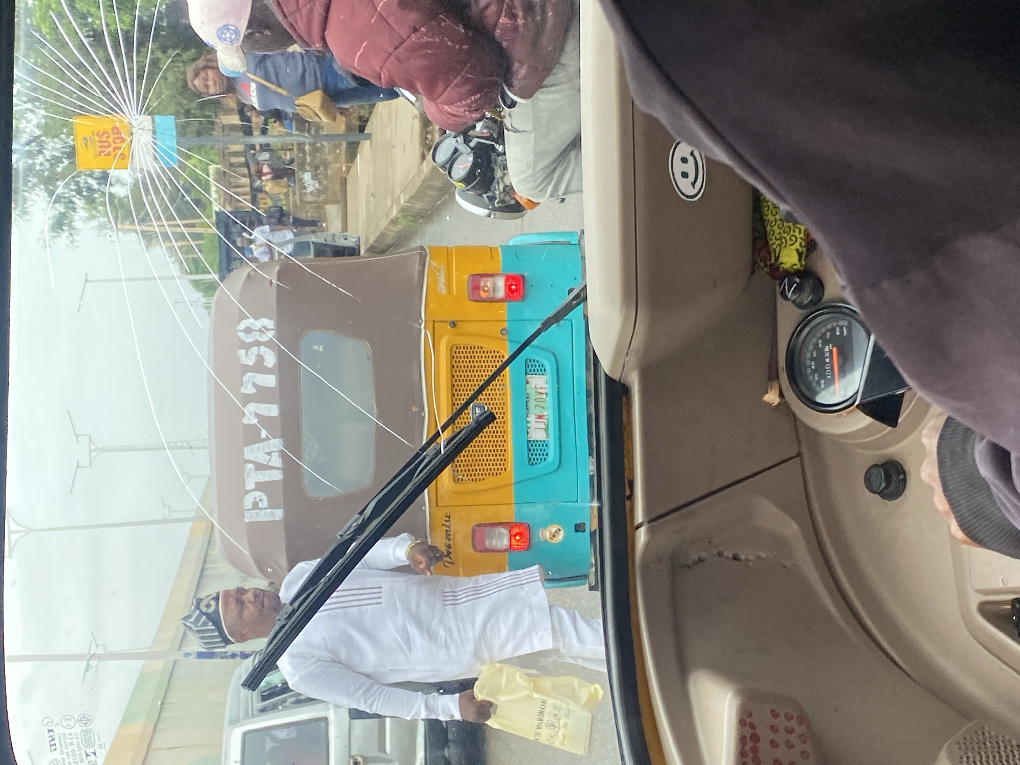 The image is taken from inside a vehicle, looking through a cracked windshield. The scene appears to be a street in Jos, Nigeria. In the foreground, a person's hands are visible on the steering wheel of the vehicle. A smiley face sticker is affixed to the dashboard.
Outside the vehicle, a man in a white traditional outfit and a cap stands holding a yellow shopping bag. He is next to a three-wheeled auto-rickshaw, which is yellow and turquoise and has "PTA 1758" written on its side. A license plate with "JJN 70YF" is visible on the rear of the auto-rickshaw. Further down the street, other vehicles and people are visible. A bus stop sign is also present. A motorcycle with a rider is parked nearby. The weather appears to be overcast.