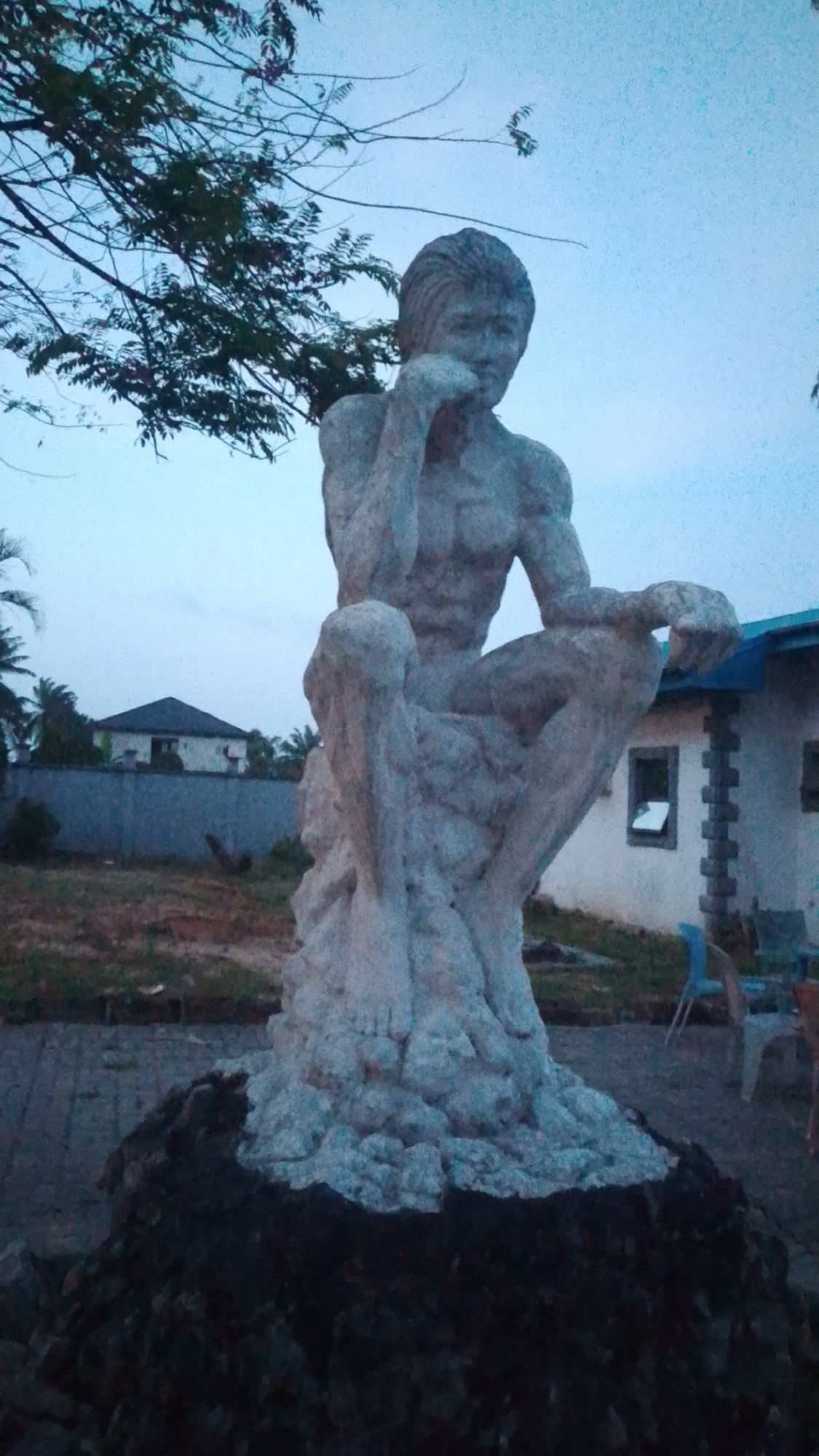The image displays an outdoor scene in Agbarho, Nigeria, featuring a prominent stone statue of a man, likely at dusk or dawn given the soft, diffused light and pale blue-grey sky. The statue depicts a muscular, shirtless male figure in a contemplative pose, seated or crouched with his right elbow resting on his knee and his chin supported by his hand. Its rough, textured surface suggests it is carved from stone or concrete.
The statue rests on a dark, rocky base, which transitions into a lighter, sculpted foundation beneath the figure. In the upper left quadrant, branches of a tree with sparse foliage extend into the sky. The background reveals a semi-developed area with residential buildings and natural elements. To the left, a white house with a dark roof is visible behind a low wall, surrounded by palm trees and other vegetation. To the right, another building with a light facade and a blue roof features an open window, with a few plastic chairs (one blue, one brownish) scattered nearby on what appears to be a dirt or grassy area. The ground immediately around the statue's base seems paved. No visible text is present. The overall atmosphere suggests quiet contemplation in a transitional light setting.