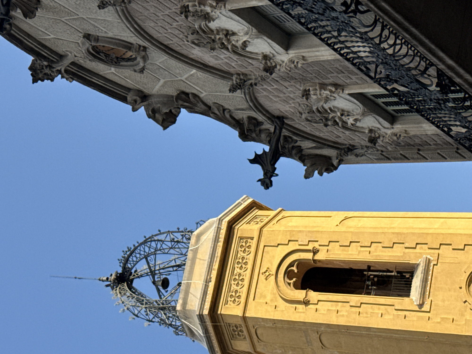 The image shows an upward view of architectural details against a clear blue sky. At the top is a section of a building with ornate stonework, including gargoyles and decorative carvings. Ironwork railings are visible on a balcony. Below, there is a tower-like structure with a yellow facade, decorated with patterned carvings. A window opening reveals what appears to be a bell inside. Beside the tower, a skeletal metal structure resembling a tower with a bell and antenna extends upwards. This is likely the Torre de les Aigües del Besòs, an element of the 1929 Barcelona International Exposition, standing near the Agbar Tower.