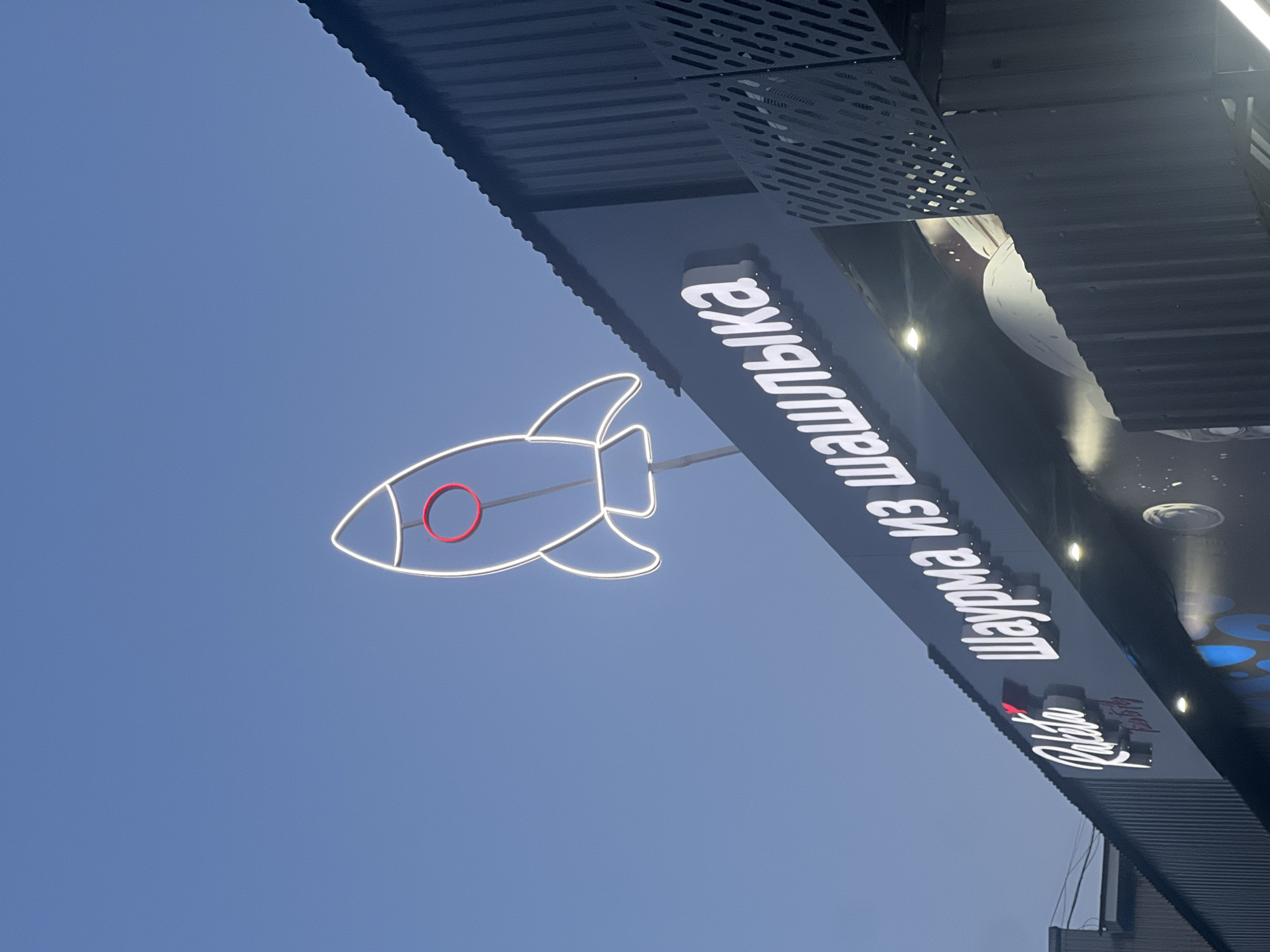 The image depicts a storefront in Chaikovsky, Russia. A large sign displays the Russian words "Шаурма из шашлыка," which translates to "Shawarma from shashlik." A neon sign of a white rocket with a red circle inside the nose is mounted on the sign. The building has a dark-colored, corrugated metal roof. The sky is light blue. Below the main sign, a smaller sign with what appears to be the name "Rocketto" is visible.