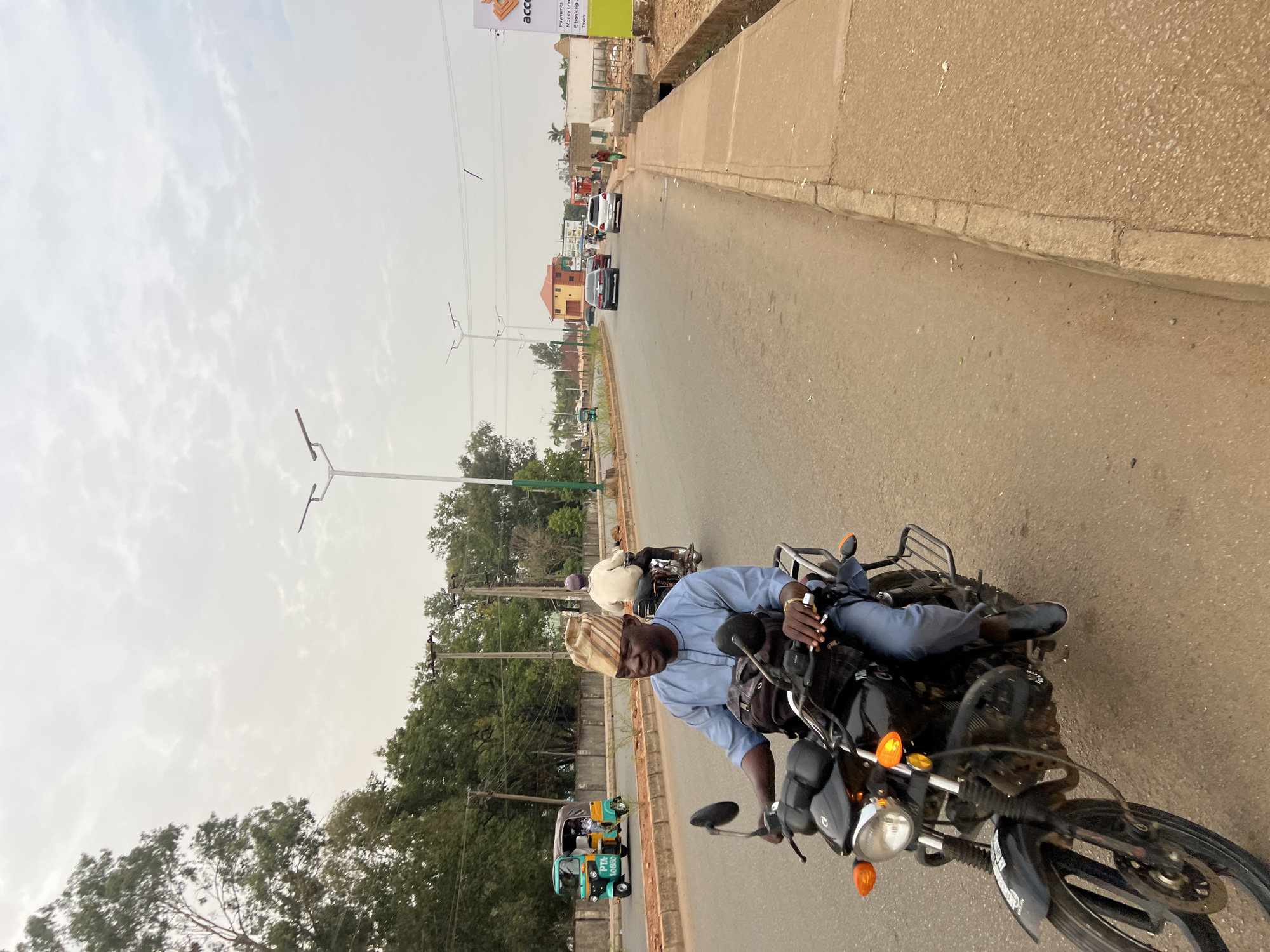 A man wearing a striped cap and a blue shirt is seated on a black motorcycle on a paved road in Bukuru, Nigeria. He is holding a cigarette in his hand. Another motorcycle is visible ahead of him, and a tuk-tuk is on the side of the road. The road is lined with trees and street lights. Cars are visible in the distance. The sky is overcast.