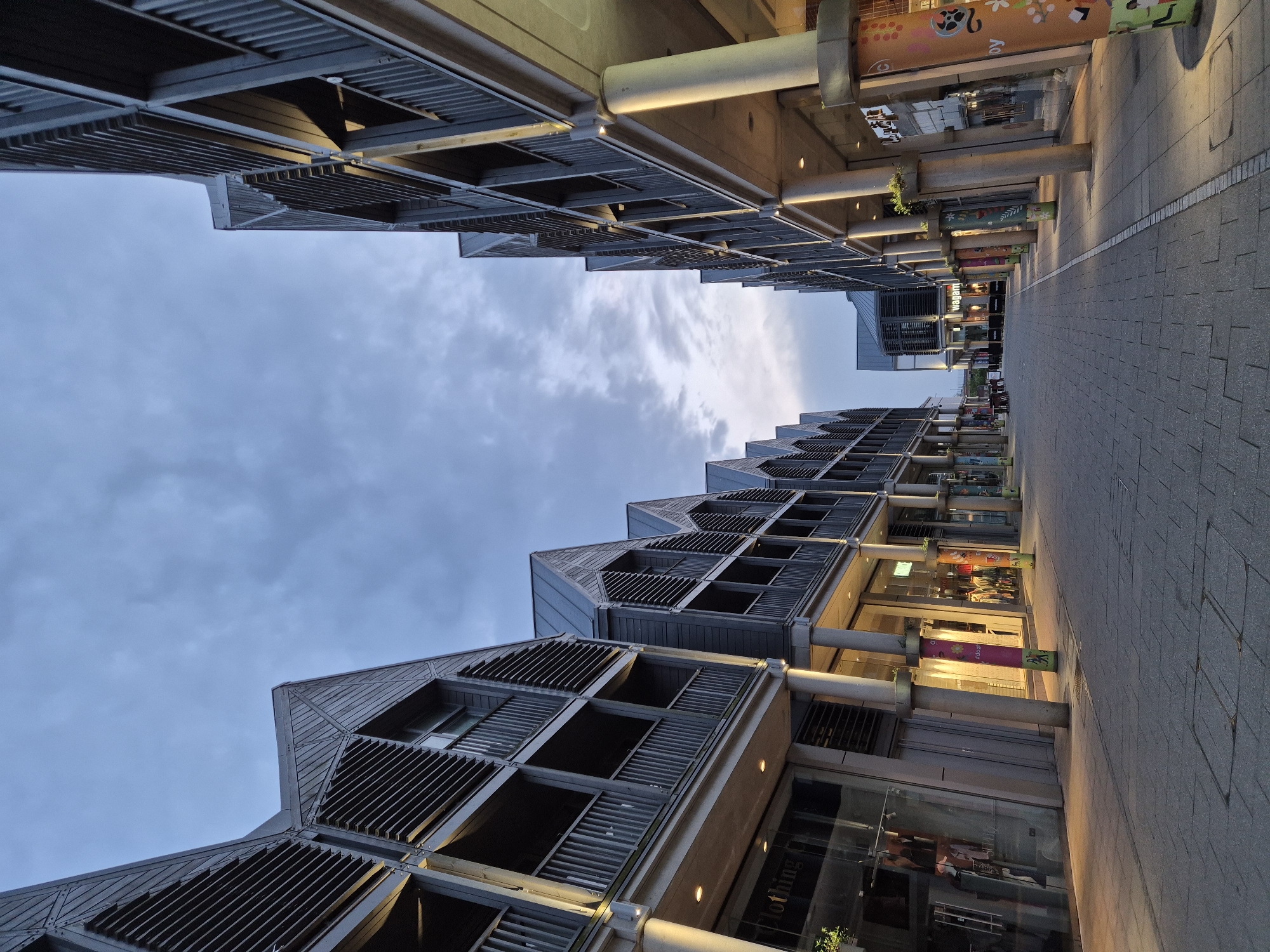 A modern pedestrian street, likely a shopping or commercial area, is depicted under an overcast sky in West Suffolk, United Kingdom. The image, originally rotated, shows two multi-story buildings flanking a paved walkway.
The architecture of the buildings is distinctive and contemporary, featuring dark grey facades with angular rooflines and prominent louvered sections, possibly for sun shading or aesthetic appeal. The ground floors of these buildings are lined with large glass windows, indicating retail stores or businesses. Recessed lighting illuminates the undersides of the building overhangs and the shop interiors, suggesting it is either late afternoon, early evening, or a generally dim day.
The walkway itself is paved with light grey rectangular tiles, and a lighter strip runs down its center. Concrete support columns line the pathway, extending upwards to the upper levels of the buildings. Some of these columns are adorned with colorful, graphic wraps.
Visible text on the column wraps includes "Wagamama," identifying a Japanese restaurant chain. Another partially visible word on a column appears to end in "py," possibly part of "Happy" or "Enjoy," surrounded by playful illustrations. On a shop window, text like "Clothing Co." or "Clothing Company" can be discerned. The sky overhead is cloudy, with soft, diffused light, yet no direct sunlight is apparent. There are no people visible in the scene, contributing to a quiet, serene atmosphere.
