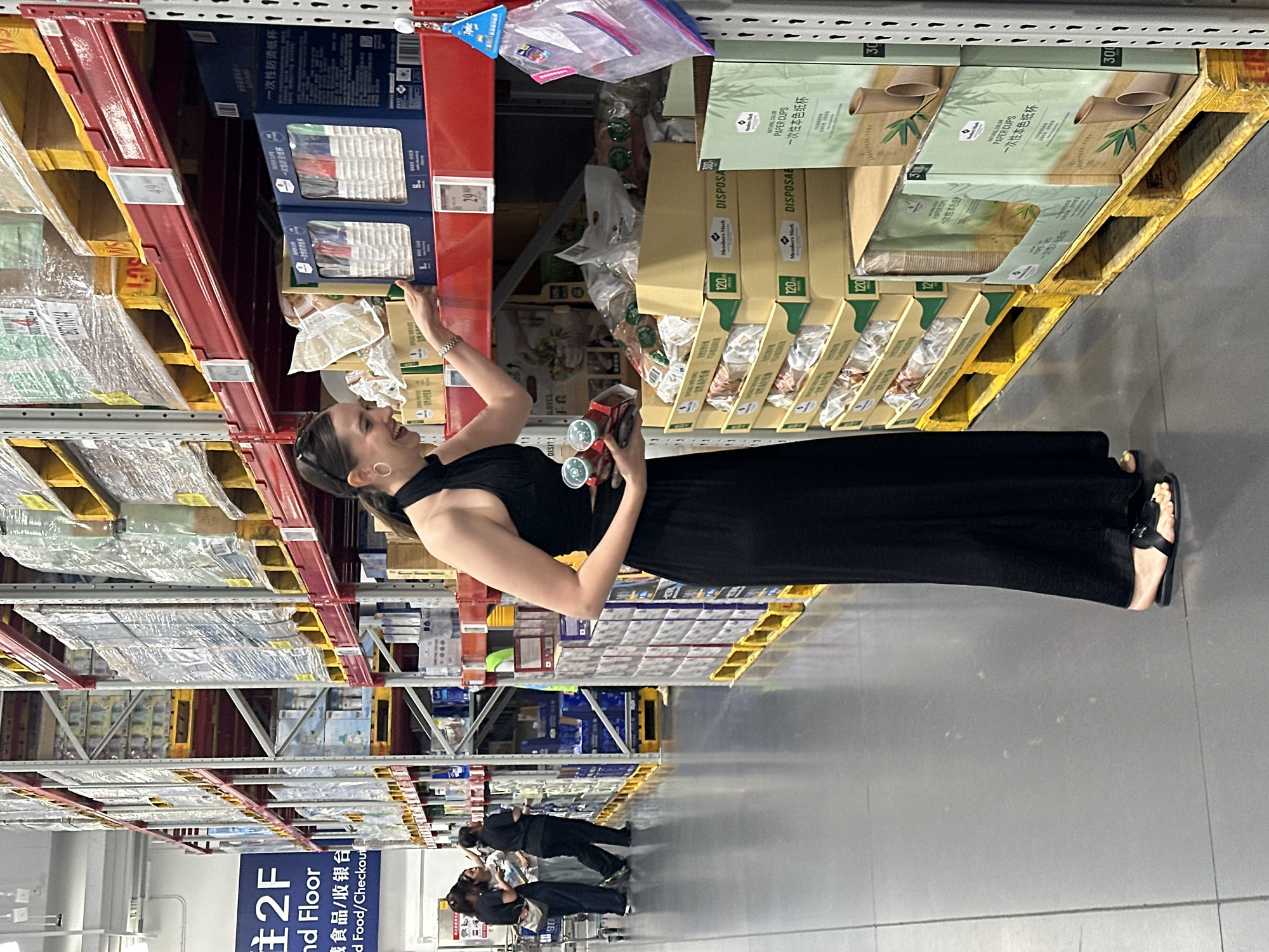 The image depicts a woman in a black halter dress and black sandals standing in an aisle of a warehouse-style store, possibly a Sam's Club, located in the Liwan District, China. She is holding two small, red-capped cylindrical objects in her left hand and reaching out with her right hand to touch a box on a shelf. The shelves are stacked high with various products, predominantly packaged goods. In the background, three other individuals, two women and one person in a black uniform, are standing near a shopping cart, engaged in conversation. A sign in the background indicates "2F" and "Food/Checkout." The floor is a polished gray surface, and the lighting appears to be overhead fluorescent.