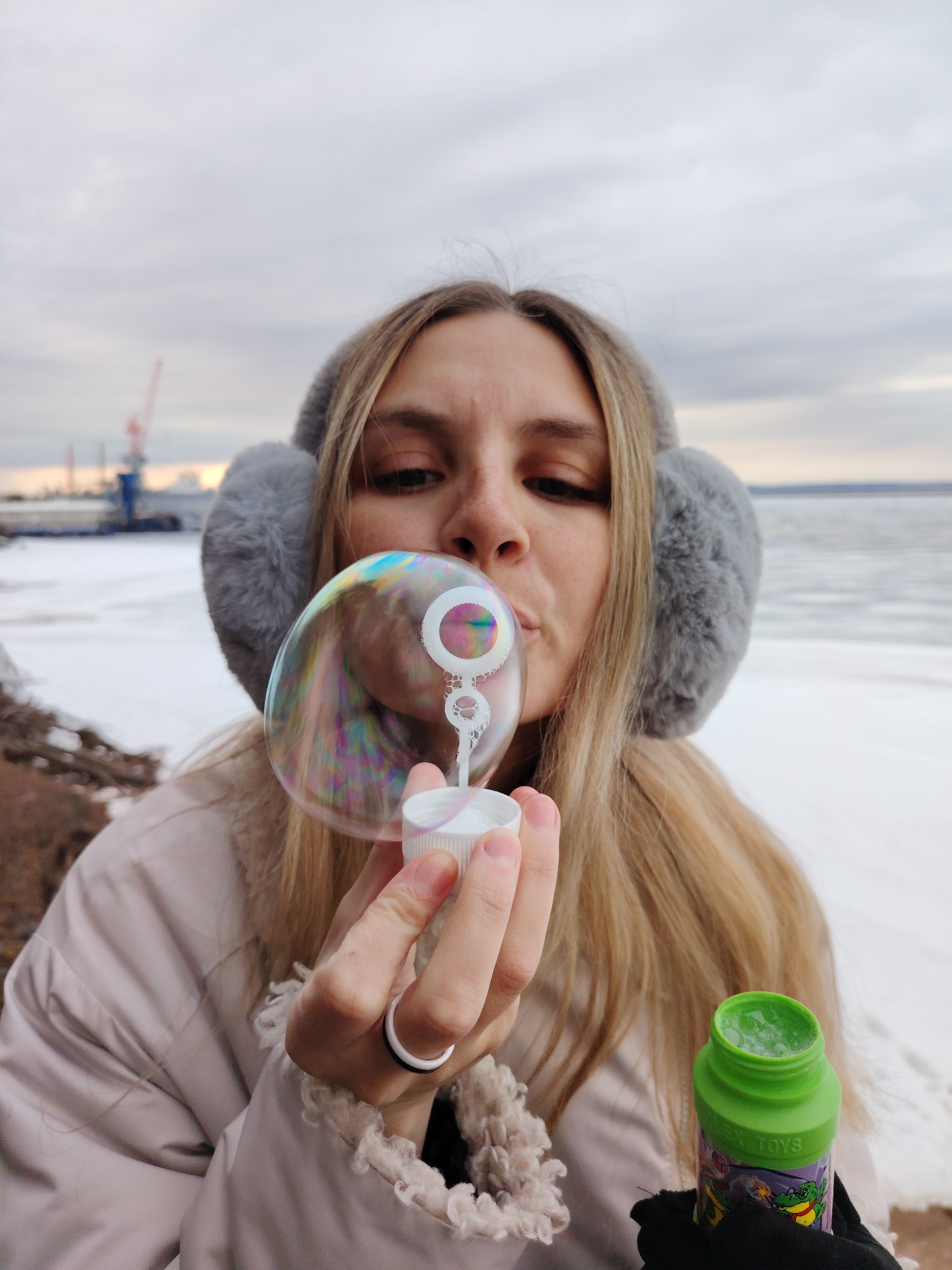 A woman with long blonde hair and gray earmuffs is blowing bubbles outdoors. She is holding a white bubble wand with a large iridescent bubble forming at the end. A green bottle of bubble solution is visible in her hand. The background shows a gray, overcast sky and a body of water with ice or snow along the shore. A crane and industrial structures are visible in the distance. The location is Chaikovsky, Russia.