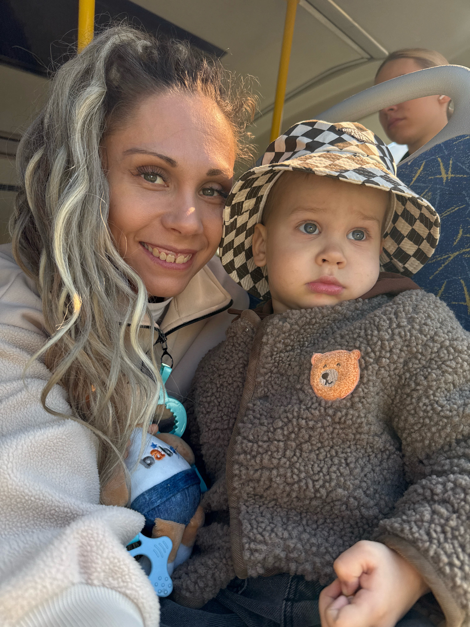 In Rusanovo, Russia, a woman with long, wavy blonde hair and light green eyes smiles at the camera while holding a young child. The child, wearing a bucket hat with a checkered pattern and a fuzzy brown jacket with a bear applique, looks slightly to the side with a neutral expression. A blue baby toy is visible between them. In the background, another person's head is partially visible, seated in what appears to be a public transport vehicle, indicated by the yellow pole and blue patterned seat. The lighting suggests it is daytime, possibly sunny.