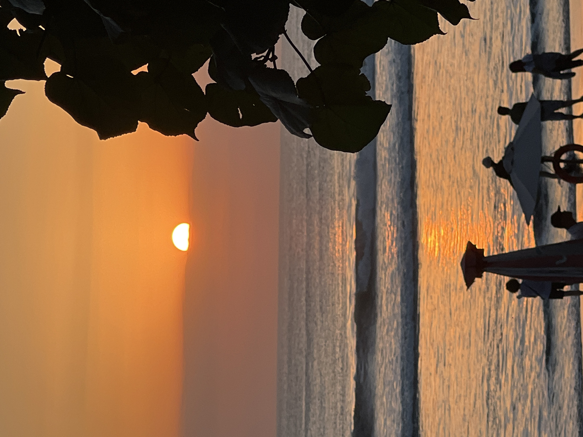 The image depicts a sunset at Kuta Beach, Indonesia. The sun, a bright orange orb partially obscured by a hazy horizon, casts a warm glow across the sky and water. Waves gently lap the shore, reflecting the sunset's colors. In the foreground, silhouetted figures of people are visible on the beach, some standing near beach umbrellas and a red and white striped object that might be a beach umbrella or a stand. Dark foliage from a tree drapes down from the right side of the frame, framing the scene.
